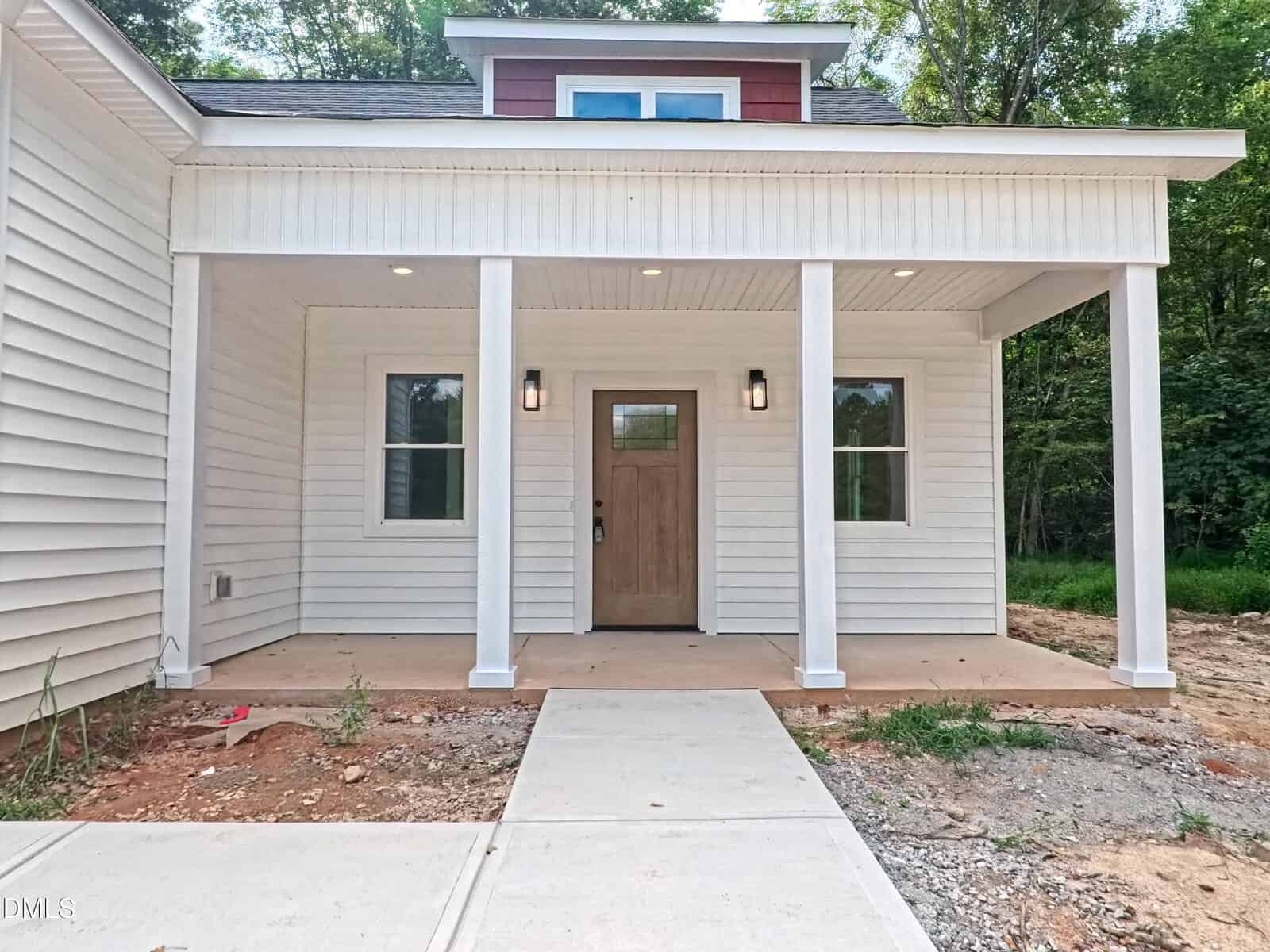Front porch of a modern house with white siding, four white pillars, a wooden front door with glass, two windows, and overhead lights; surrounded by trees and unfinished landscaping.