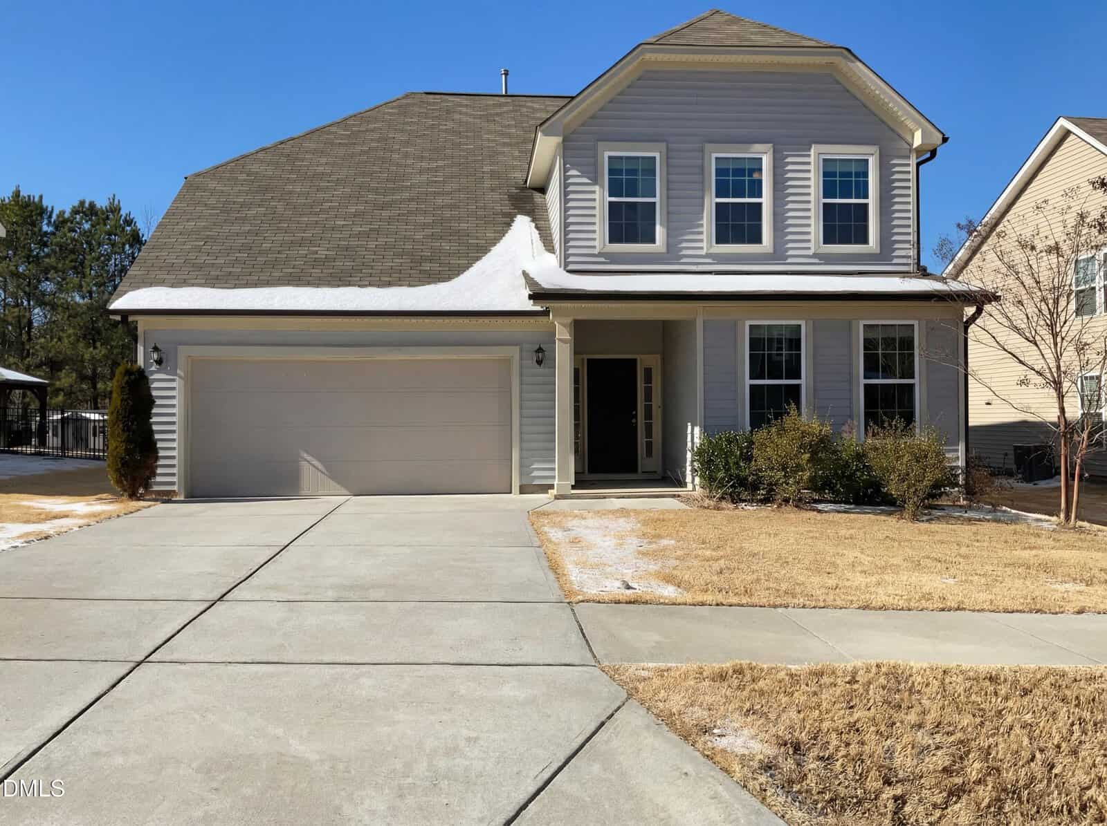 Two-story suburban house with gray siding, attached two-car garage, and a covered front porch. The driveway and lawn have patches of snow, and there are shrubs along the front of the house.