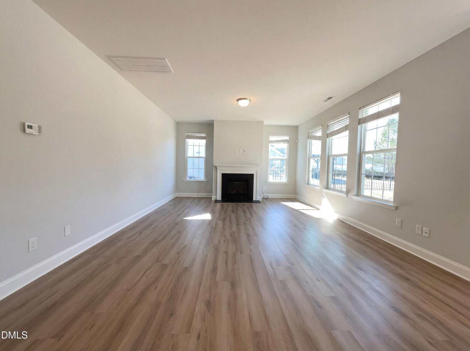Bright, empty living room with light wood floors, large windows letting in natural light, a white fireplace centered on the far wall, and neutral-colored walls.