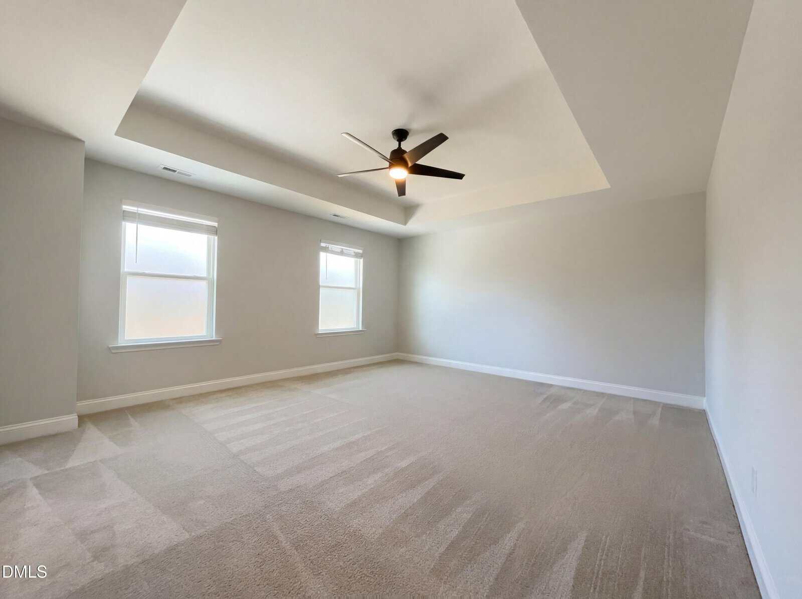 A spacious, empty room with light beige carpet, two large windows with white blinds, light-colored walls, and a modern ceiling fan with a light in the center of a tray ceiling.