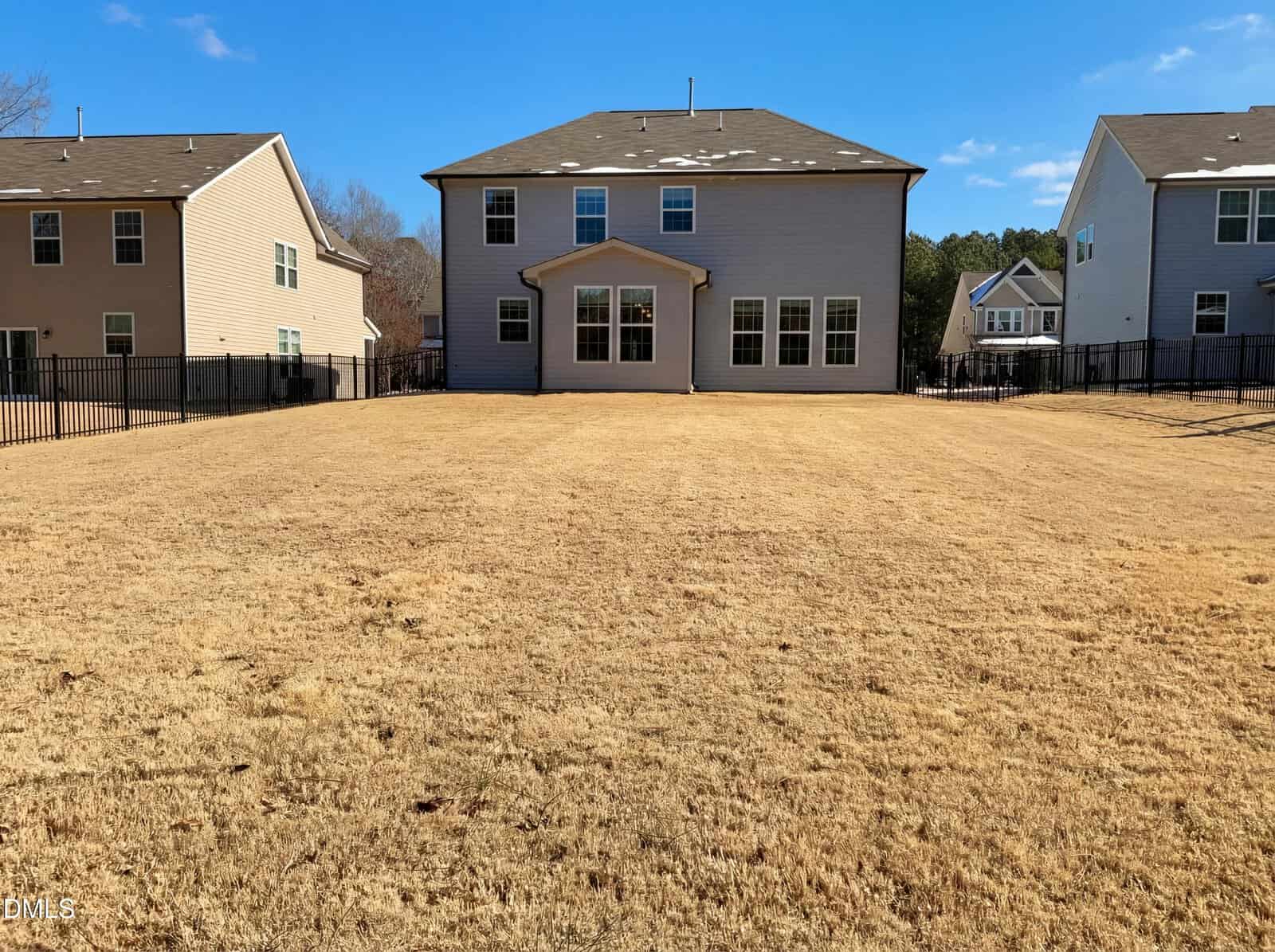 Back view of a two-story house with large windows, beige siding, and a fenced backyard with dry, yellow grass. Neighboring houses and a blue sky are visible in the background.