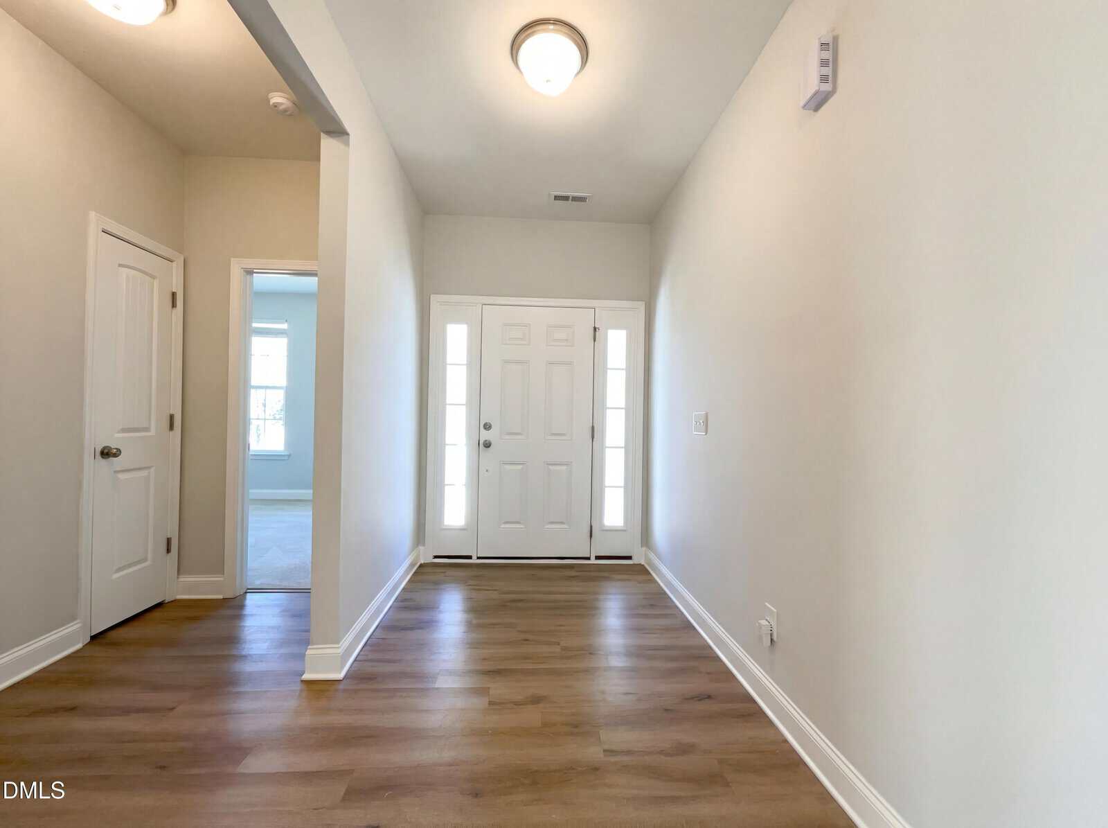 Bright entryway with wood flooring, white walls, and a white front door with sidelights. Two interior doors on the left lead to other rooms. Ceiling lights illuminate the space.