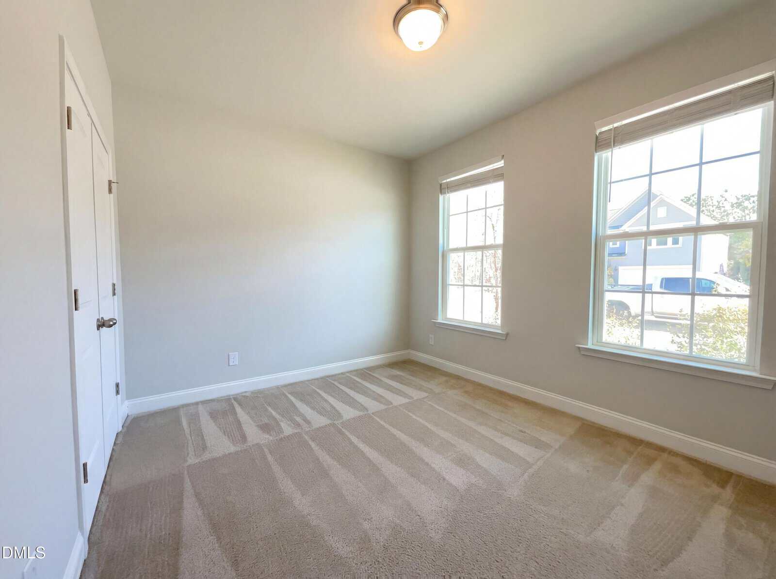 Bright, empty room with beige carpet, two large windows letting in natural light, white walls, and a closed white double door on the left. Outside the windows, part of a house and car are visible.