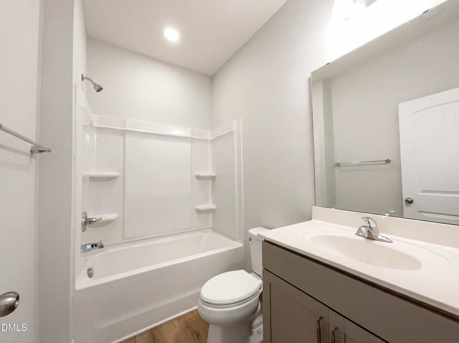Modern bathroom with a white bathtub and shower, white toilet, and a gray vanity with a sink. The room has light-colored walls, a large mirror, and wood-style flooring. Bright overhead lighting.