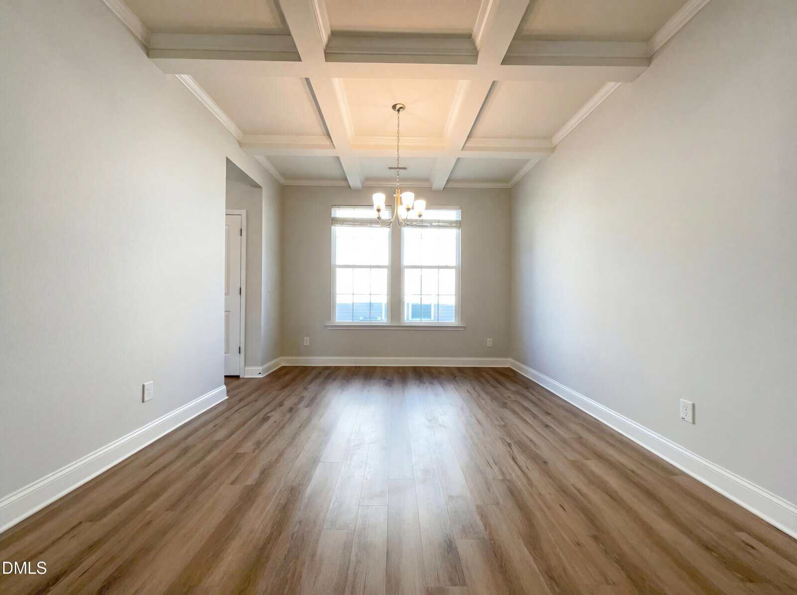 Bright, empty room with light wood flooring, coffered ceiling, and a modern chandelier centered above. Two large windows let in natural light, and walls are painted a soft neutral color.