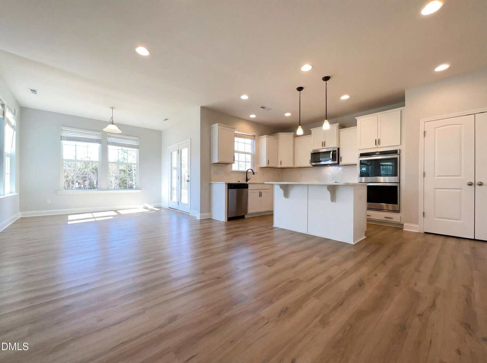 Open-concept kitchen and dining area with light wood floors, white cabinets, stainless steel appliances, an island with pendant lights, and large windows allowing natural light into the space.