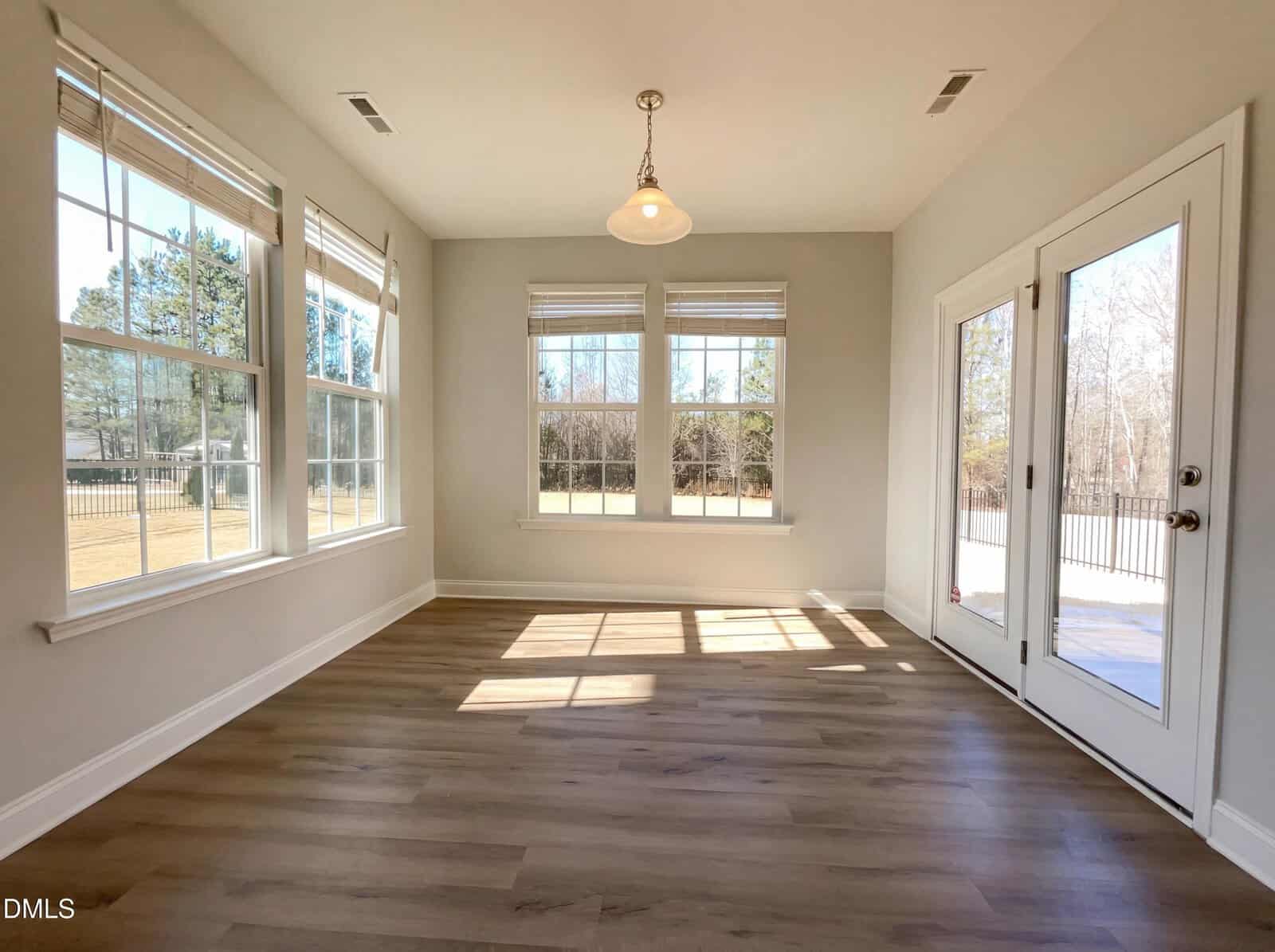 A bright, empty sunroom with large windows, light wood floors, and a glass door leading outside. Sunlight streams in, creating patterns on the floor. Trees and a lawn are visible through the windows.