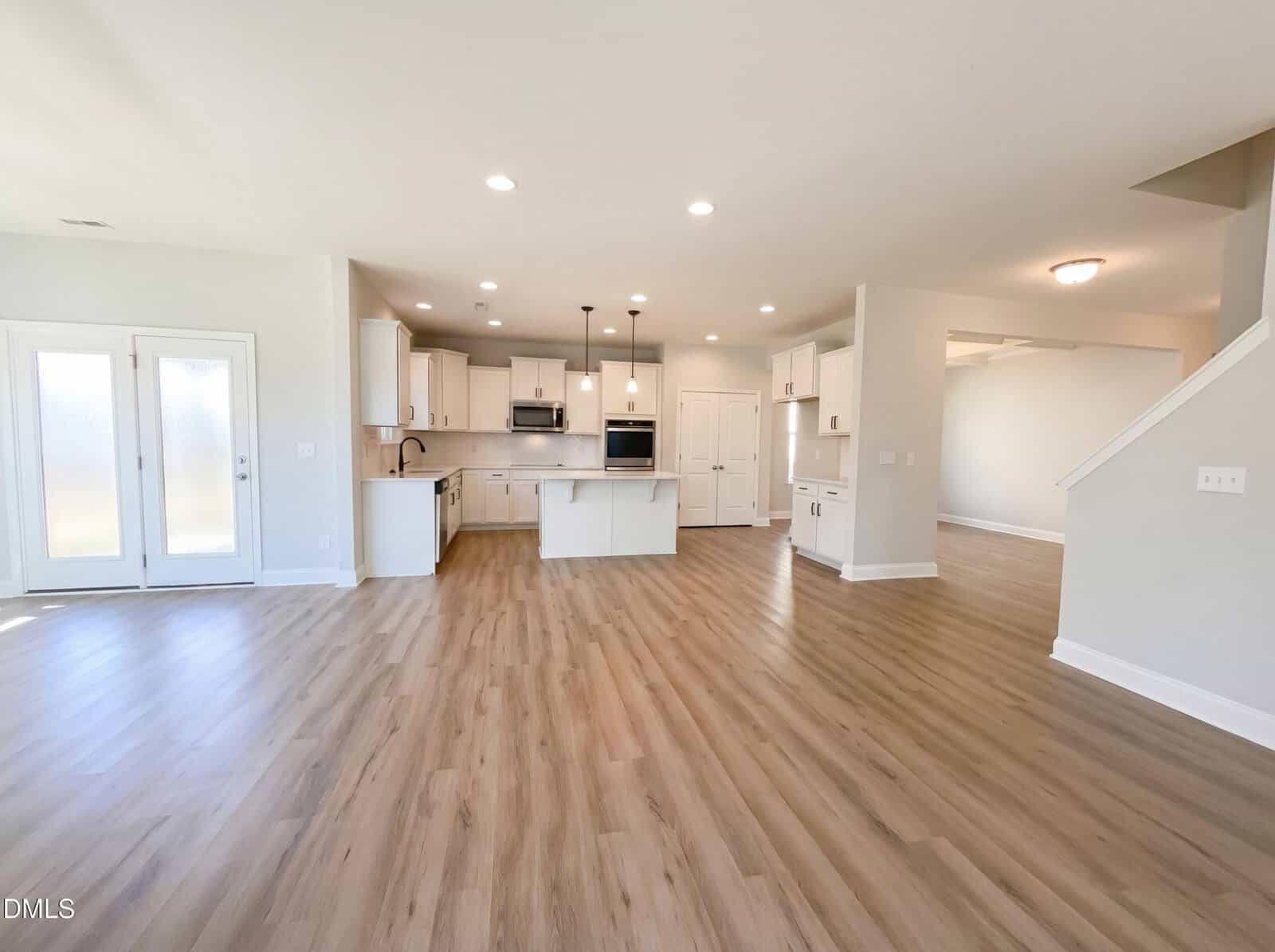 Spacious modern kitchen and living area with light wood floors, white cabinets, island, stainless steel appliances, and recessed lighting. French doors and large windows offer natural light.
