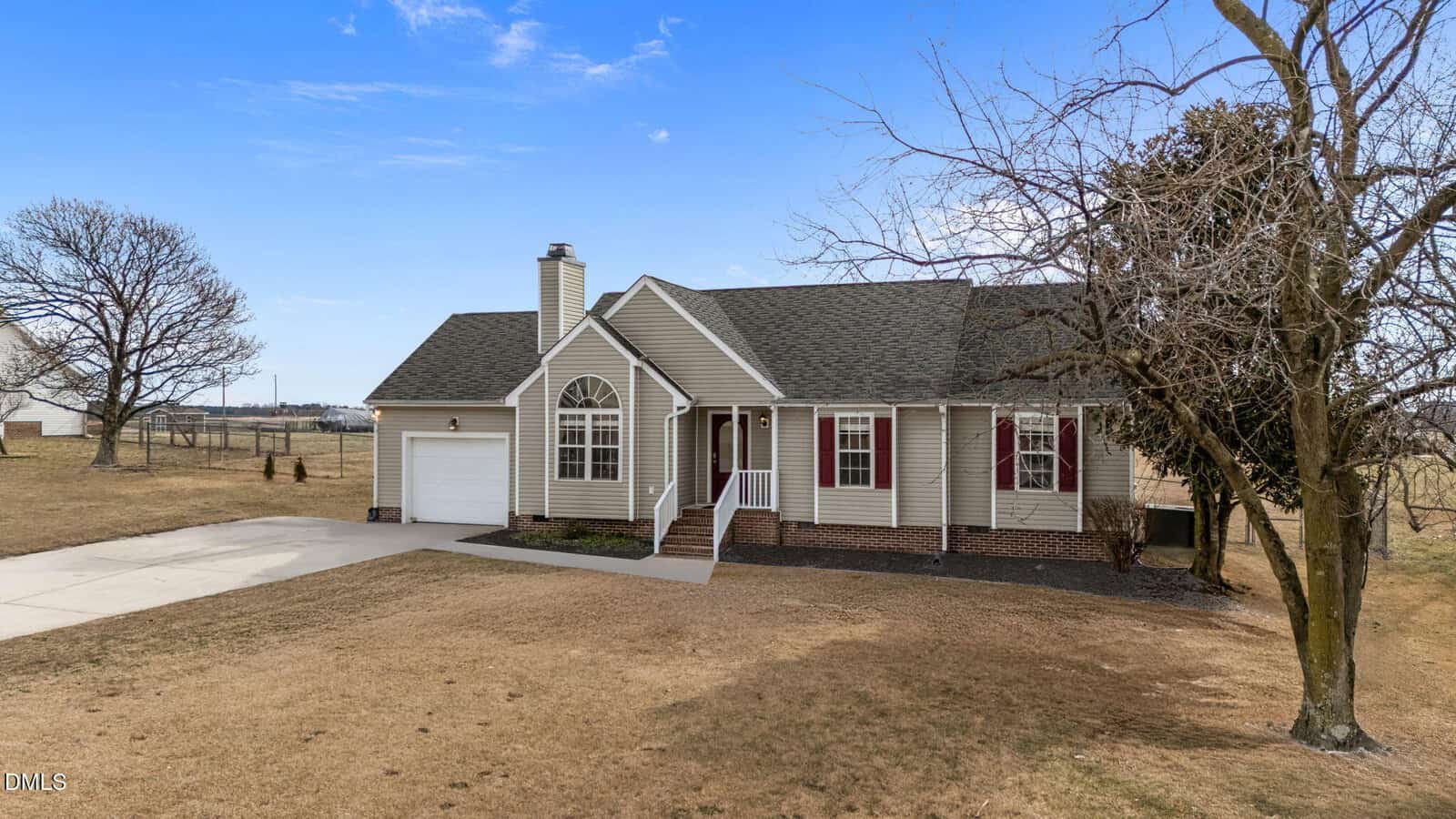 A single-story beige house with a gable roof, white trim, red front door, and attached garage sits on a large, mostly bare lawn under a clear blue sky, with a few leafless trees nearby.