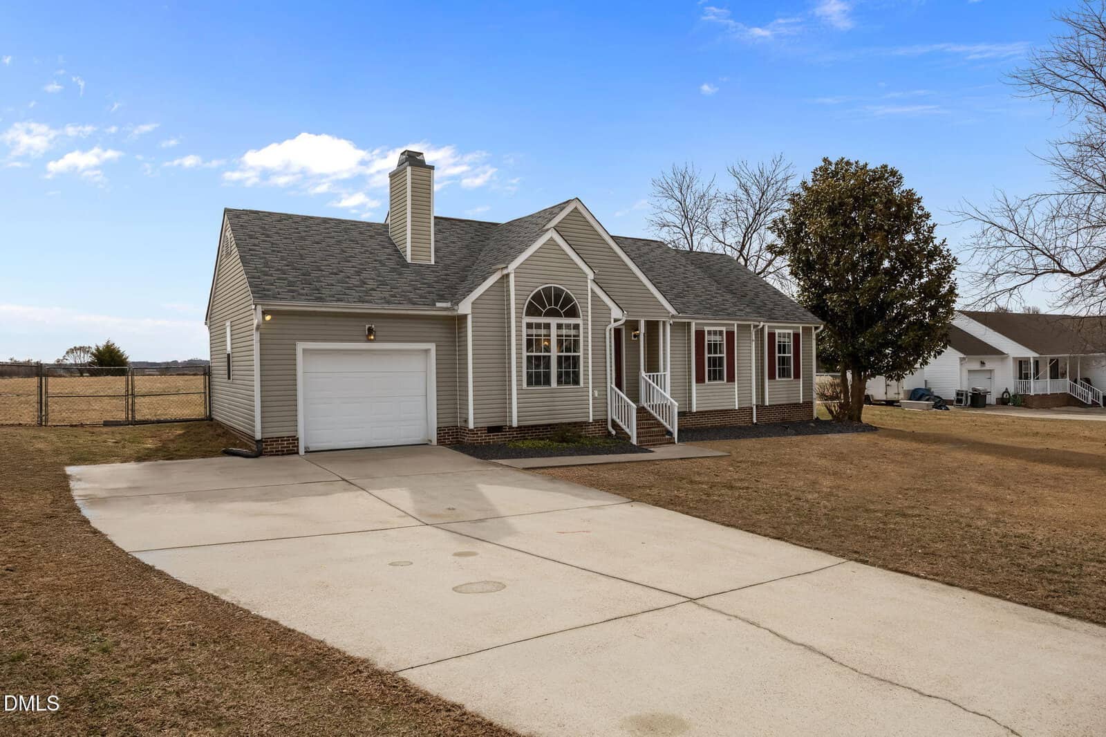 A single-story beige house with white trim, a front porch, large arched window, and a brick chimney. The house has a wide driveway, a spacious yard, and is set against a clear blue sky.
