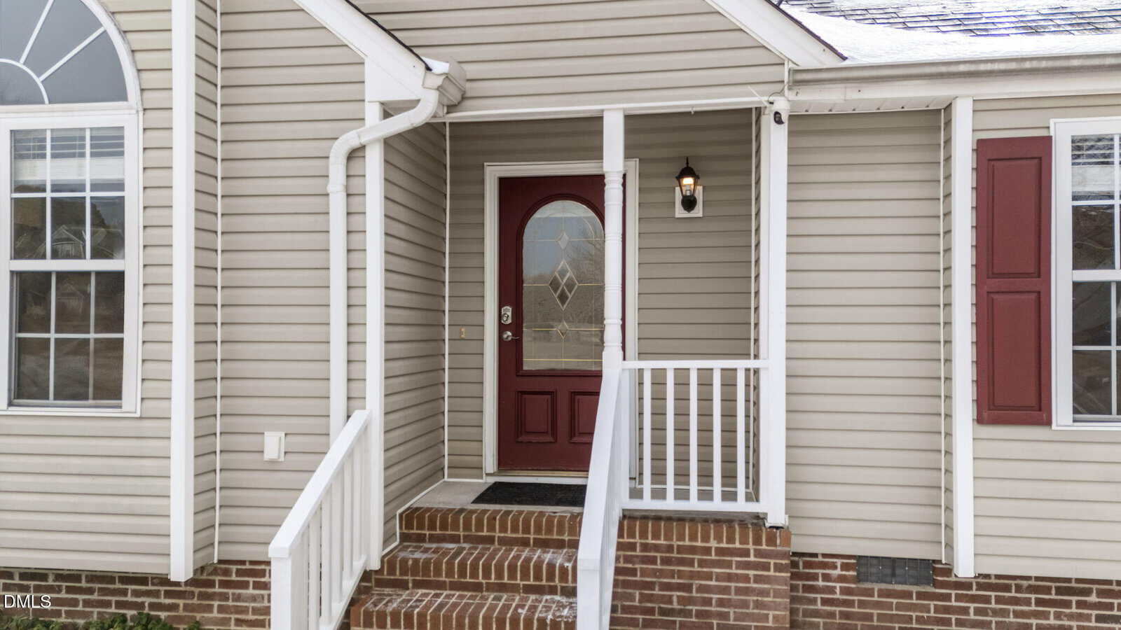 Front porch of a house with beige siding, white trim, a red door with a window, white railing, brick steps, and a wall-mounted light fixture next to the door.