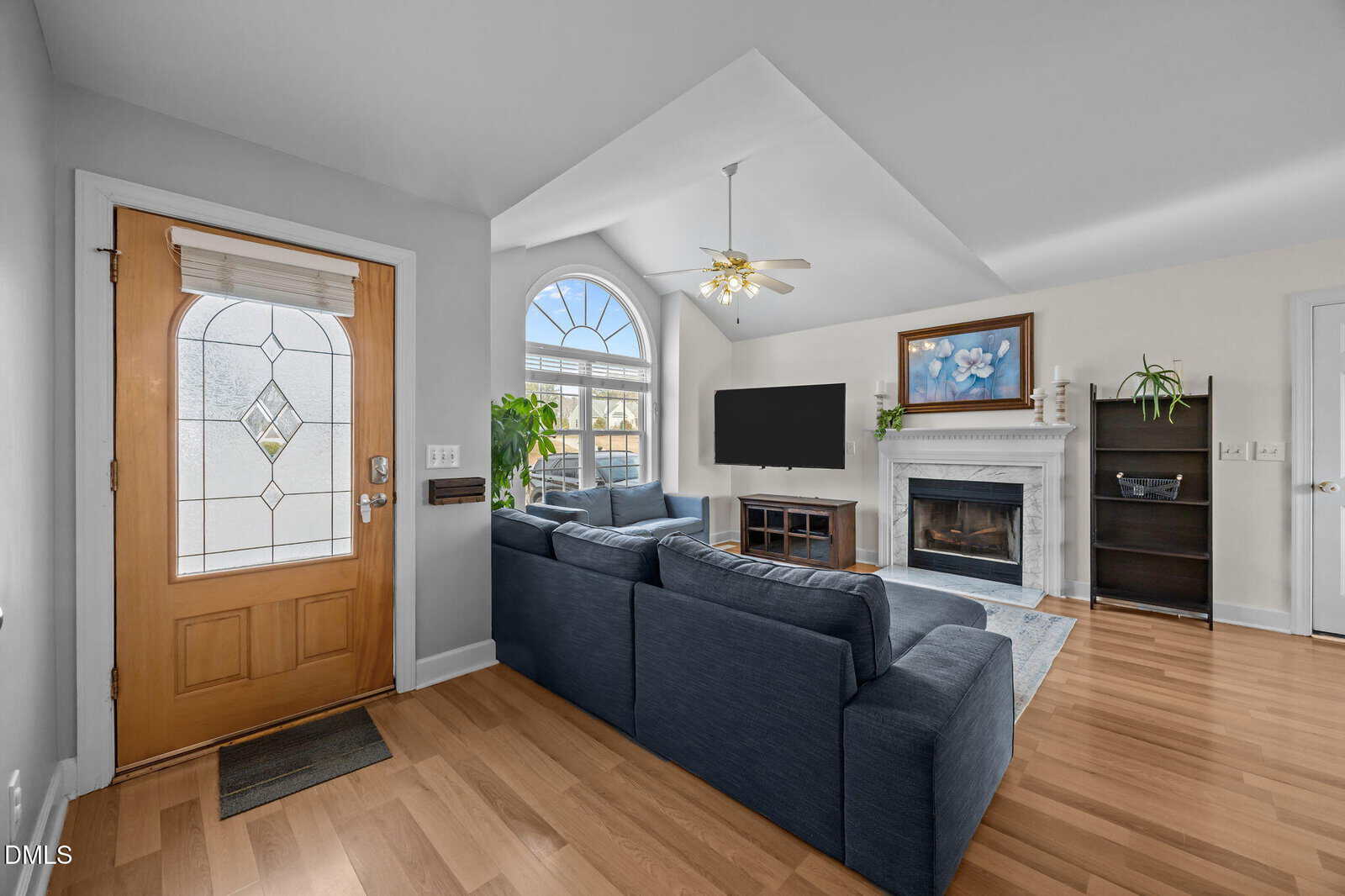 Bright living room with a vaulted ceiling, blue sectional sofa, fireplace, TV, wood flooring, a large arched window, indoor plants, and a wooden front door with a glass panel.