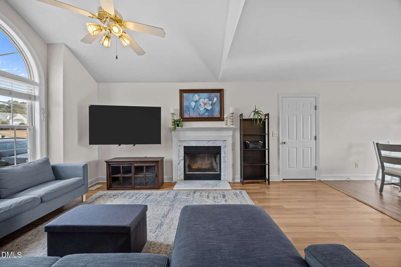 A bright living room with hardwood floors, a ceiling fan, blue sofas, a TV on a stand, a white fireplace with a floral painting above it, shelves, and a dining area in the background. Sunlight enters through a large window.
