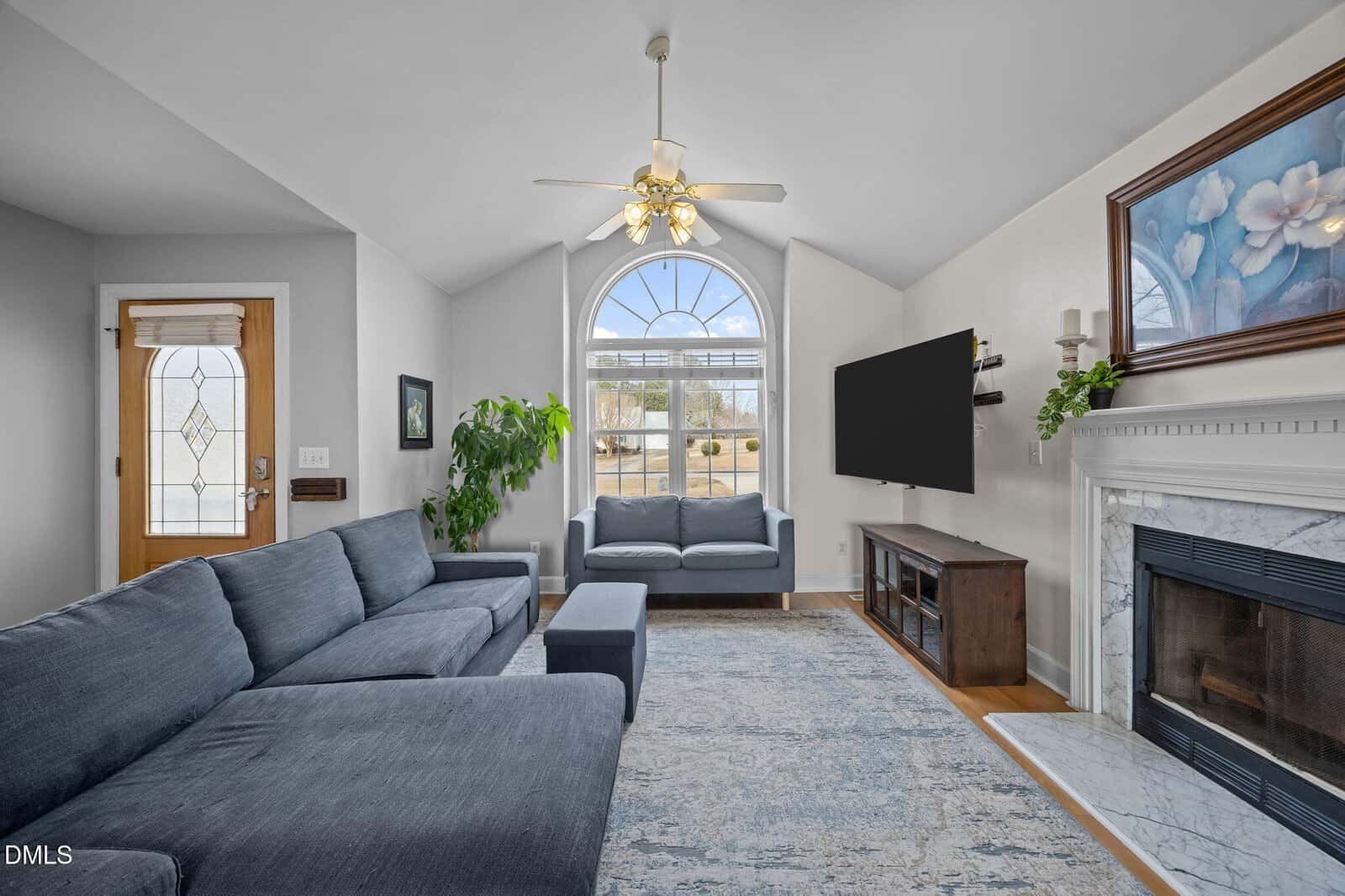 A bright living room with vaulted ceiling, large arched window, blue sectional sofa, loveseat, wall-mounted TV, fireplace with white mantel, artwork above, potted plant, and light wood floors.