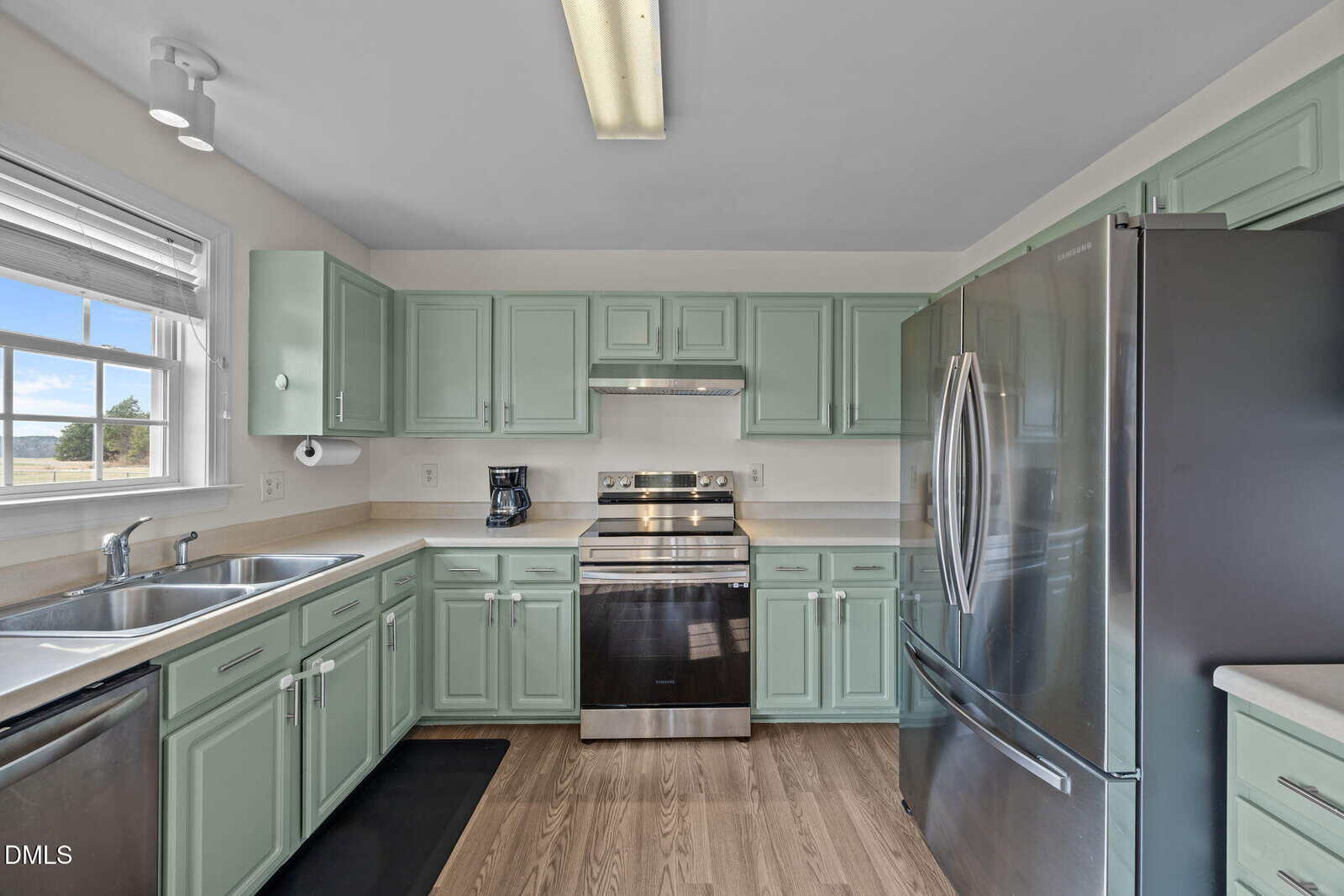 Modern kitchen with mint green cabinets, stainless steel appliances, a large double-door refrigerator, electric stove with a vent hood, double sink under a window, and light wood flooring. Natural light fills the space.