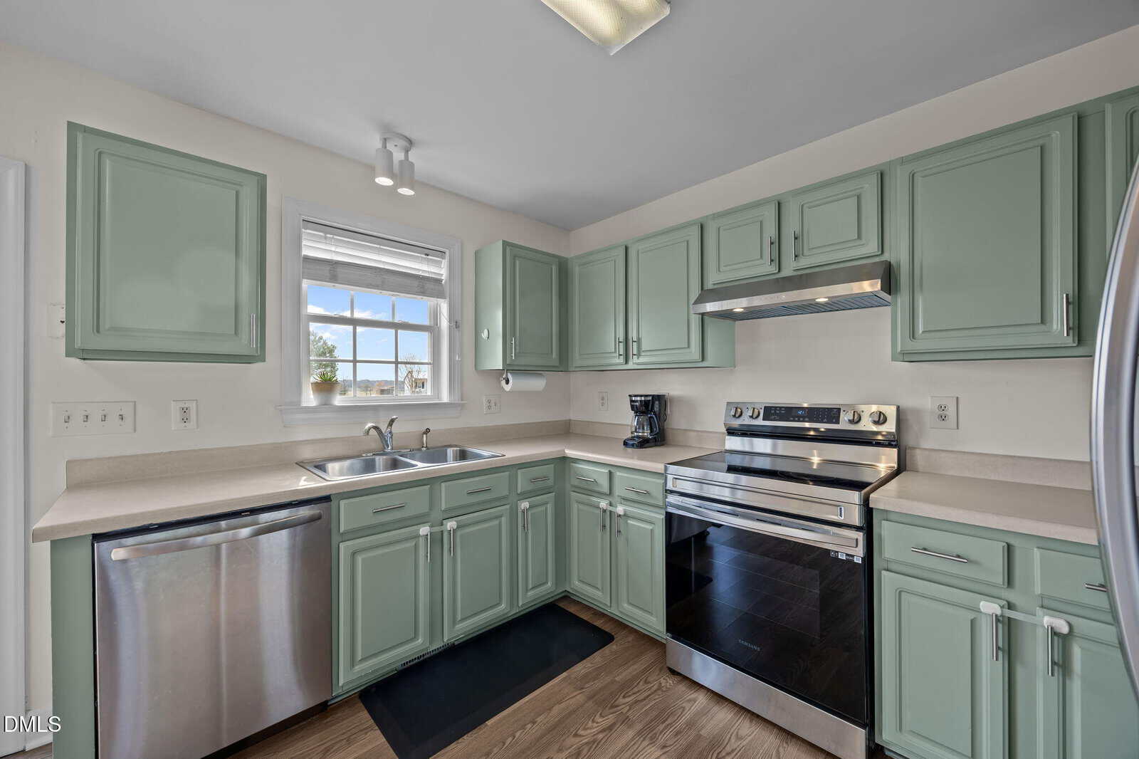A modern kitchen with mint green cabinets, stainless steel appliances, a double sink beneath a window, light countertops, and wood flooring. Natural light brightens the space.