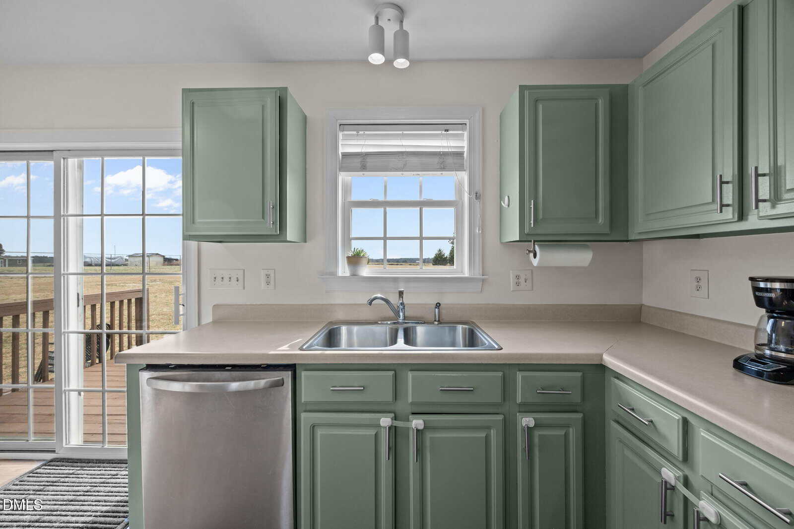 A bright kitchen with green cabinets, a double stainless steel sink under a window, beige countertops, and a stainless steel dishwasher. A sliding glass door opens to a wooden deck outside.