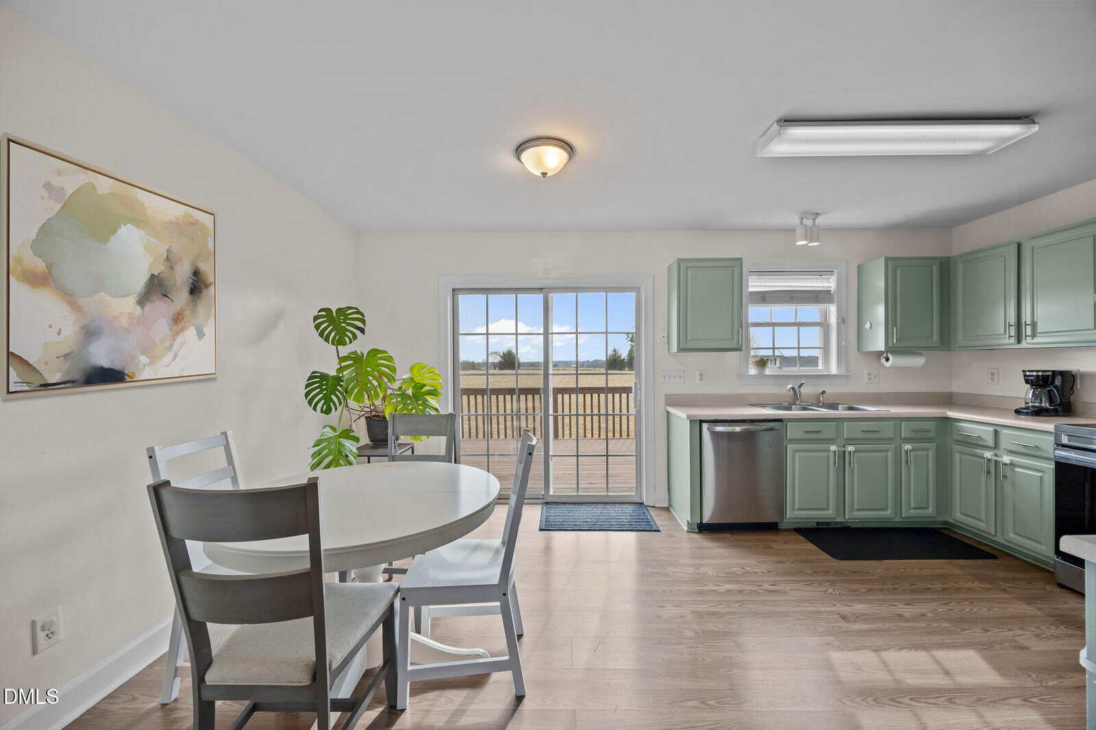 Bright kitchen and dining area with light green cabinets, round white table with four chairs, potted plant, abstract wall art, and sliding glass doors opening to a wooden deck with a view of a field.