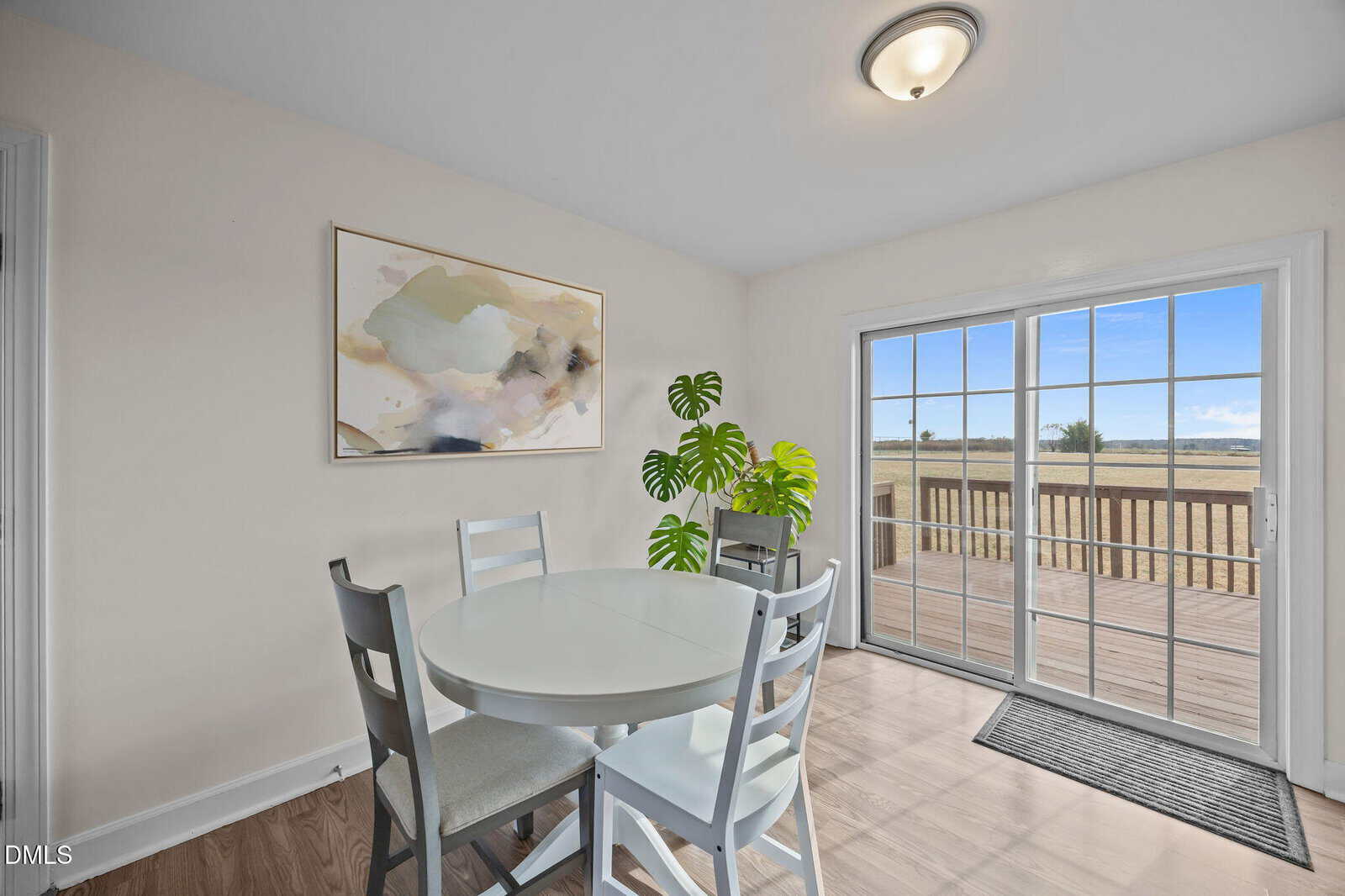 A small dining area with a round table and four chairs, a potted plant, abstract wall art, and sliding glass doors leading to a wooden deck with a view of an open field.