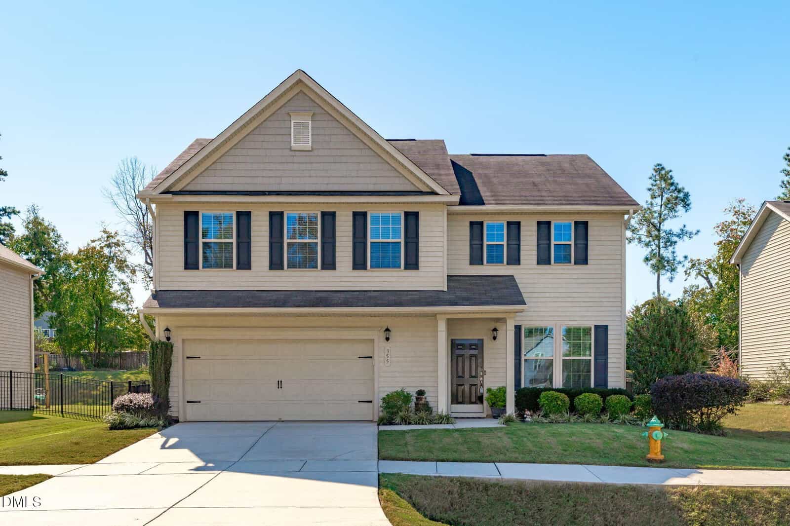 Two-story beige suburban house with black shutters, a covered front porch, double garage, and well-kept lawn. A fire hydrant is on the grass near the driveway. Trees and bushes are in the background.