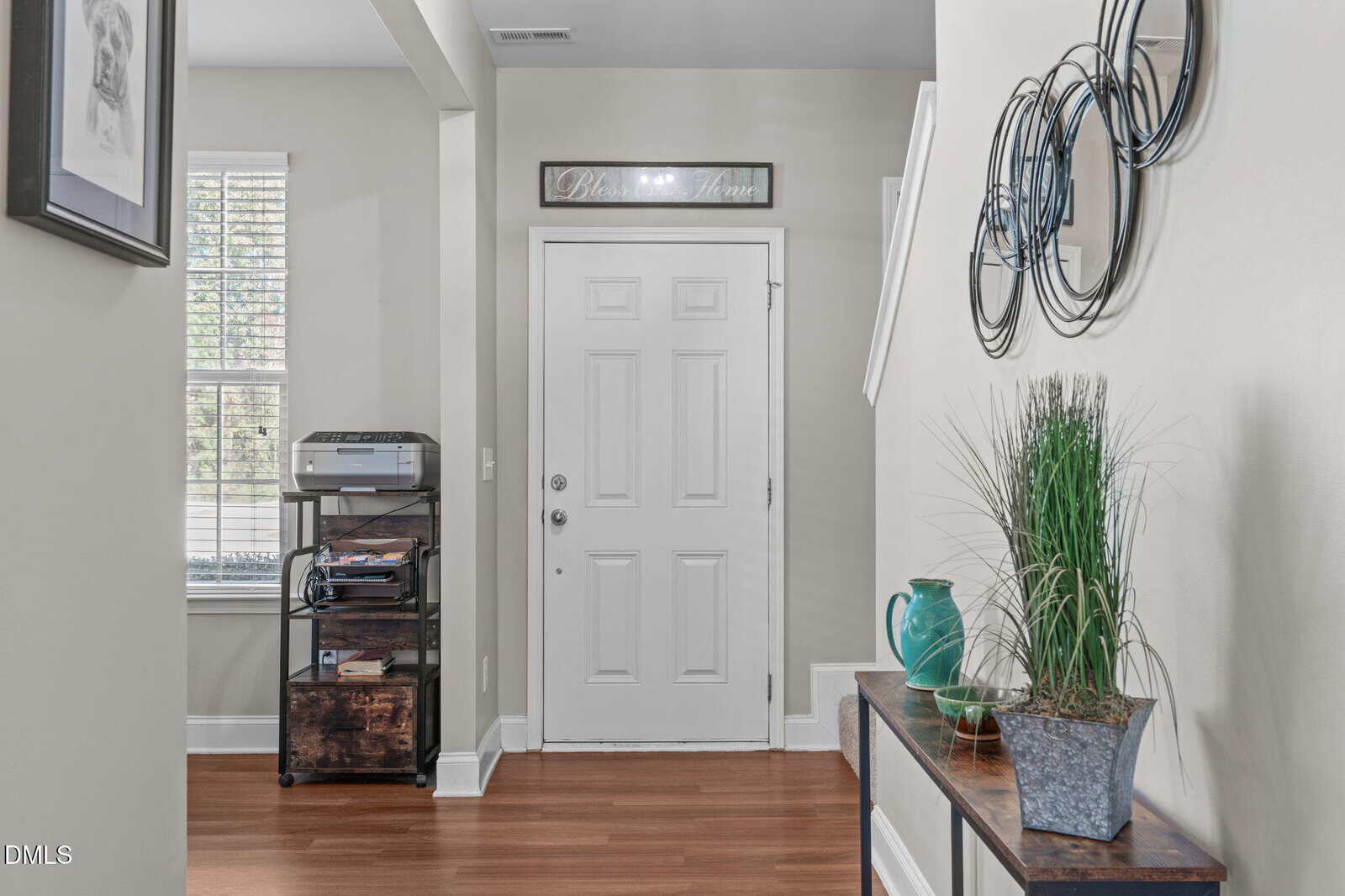 A bright entryway with a white front door, wall decor, a side table with plants and a teal vase, and a printer on a small shelf by a window with blinds. The floor is wood and the walls are light gray.