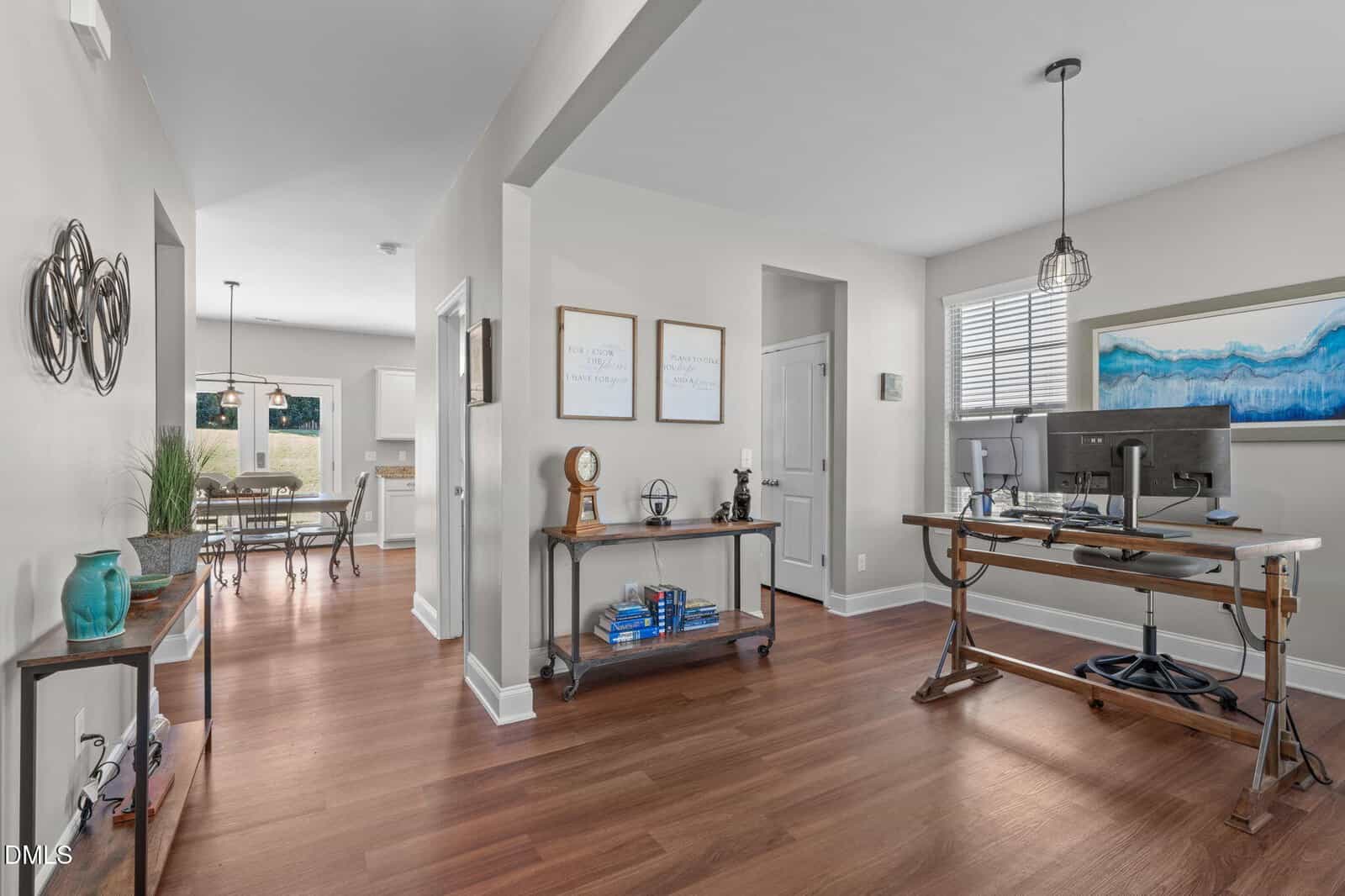 Modern home interior with a hallway leading to a dining area and kitchen, an office space with a desk and computer on the right, wooden floors, light-colored walls, and decorative art and plants throughout.