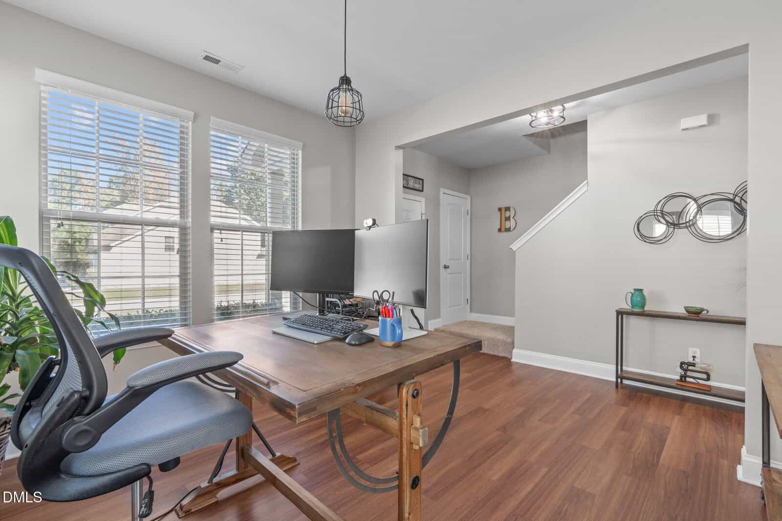 A modern home office features a large wooden desk with dual monitors, a keyboard, and office supplies. An ergonomic chair faces windows with white blinds. The room has wood floors, neutral walls, and decorative accents.