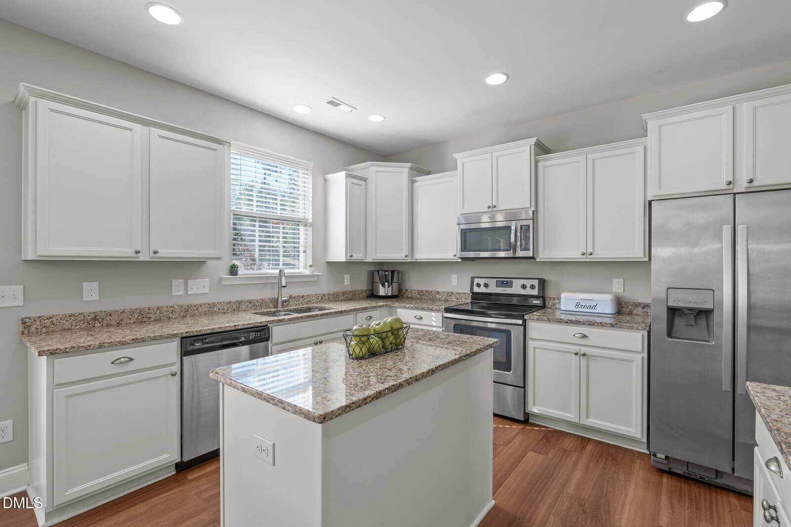 Modern kitchen with white cabinets, granite countertops, stainless steel appliances, a central island with a bowl of green apples, and wood flooring. A window above the sink lets in natural light.