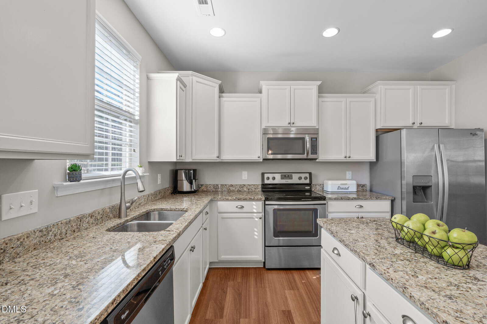 Modern kitchen with white cabinets, stainless steel appliances, granite countertops, a double sink, and a bowl of green apples on the island. Sunlight comes in through a window above the sink.