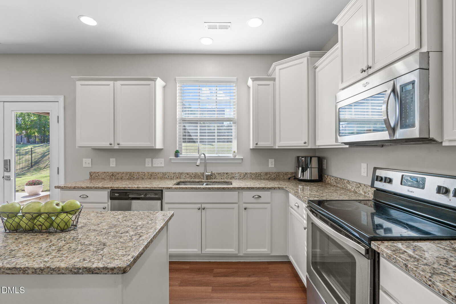Bright, modern kitchen with white cabinets, granite countertops, stainless steel appliances, a double oven, and a basket of green apples on the island. Sunlight shines through the window above the sink onto the wood floor.