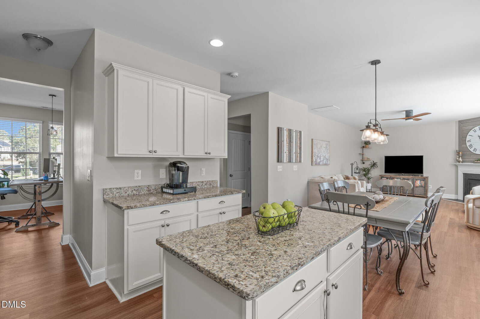 Bright, modern kitchen and dining area with white cabinets, granite countertops, a kitchen island with a bowl of green apples, wooden floors, and an open layout leading to a living room with a fireplace and wall clock.