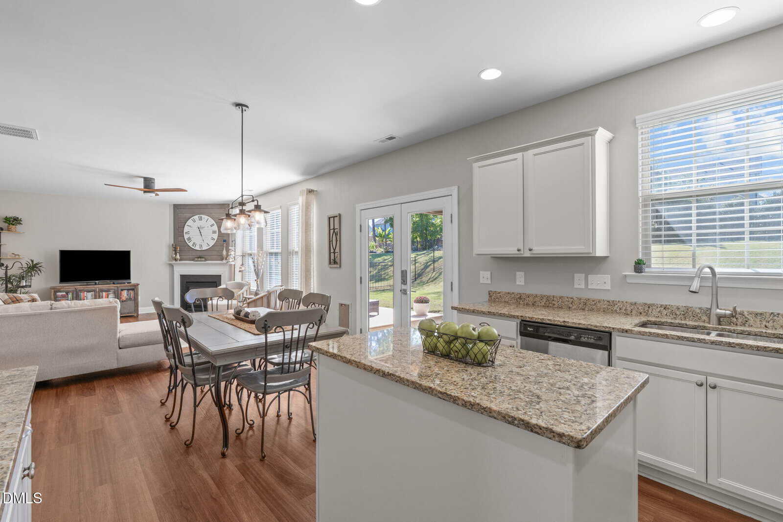 Bright kitchen and dining area with granite countertops, white cabinets, a dining table, and a cozy living room with a TV and fireplace. Large windows and glass doors let in natural light and show a green yard outside.