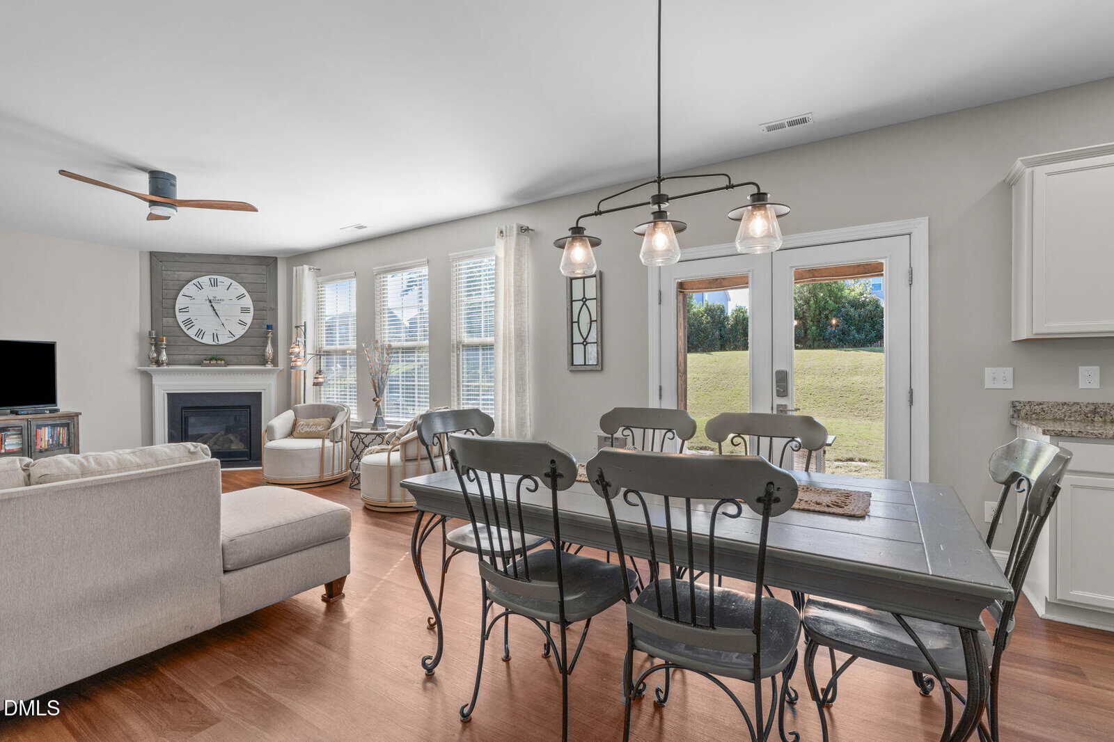 Open-concept living and dining area with a dark wood table, metal chairs, light wood floors, a sofa, a fireplace, a wall clock, large windows with curtains, and a glass door leading to a grassy backyard.