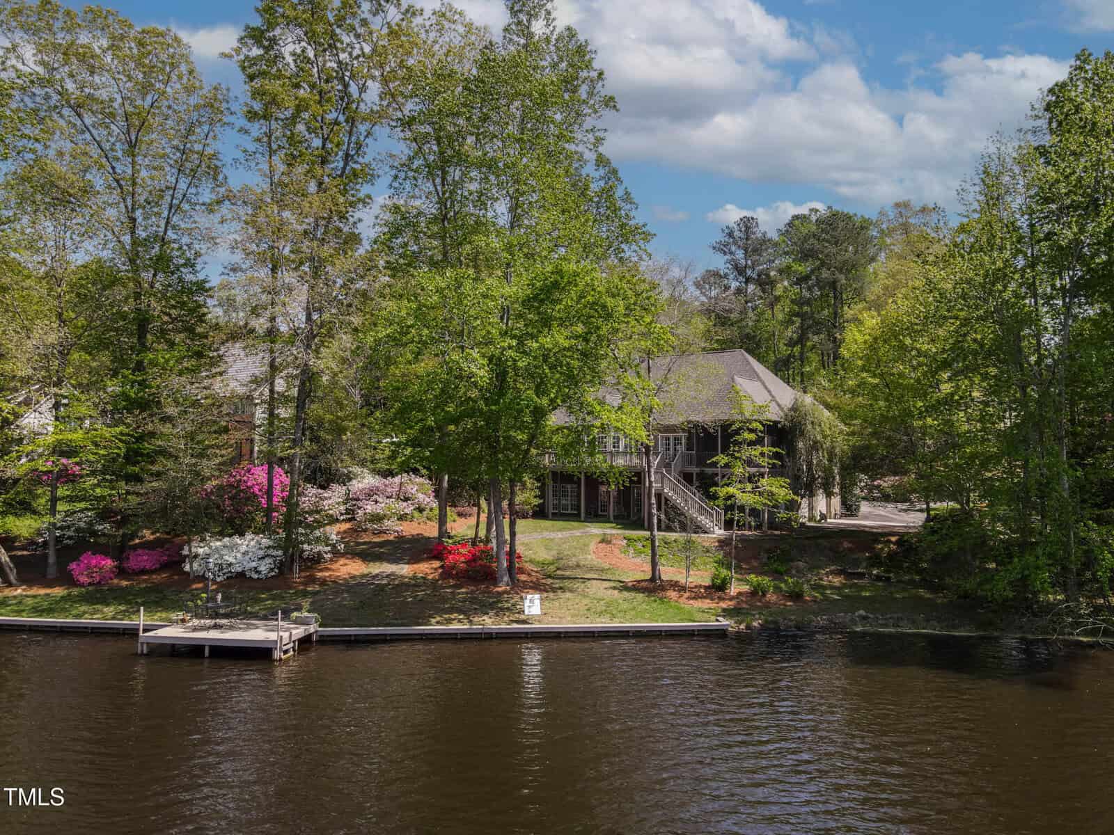 A lakeside house surrounded by tall green trees and colorful flowering bushes, with a small dock extending into the calm water under a partly cloudy sky.