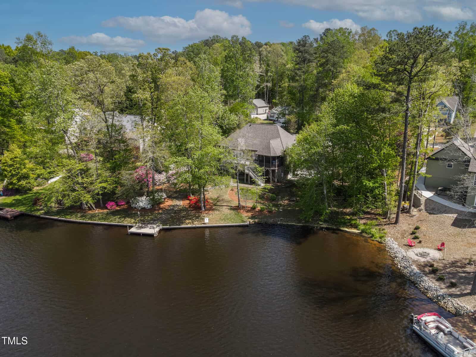 Aerial view of a lakeside house surrounded by trees, with a wooden dock extending into the water, neighboring homes nearby, and a small boat near a rocky shoreline.