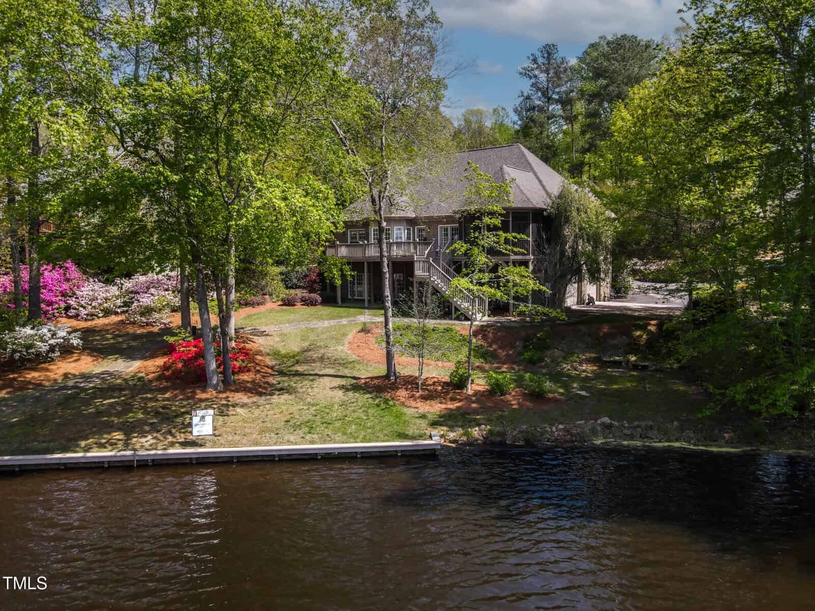 A two-story house with a sloped roof sits among tall trees and flowering bushes, overlooking a calm waterfront with a small dock in the foreground under a partly cloudy sky.