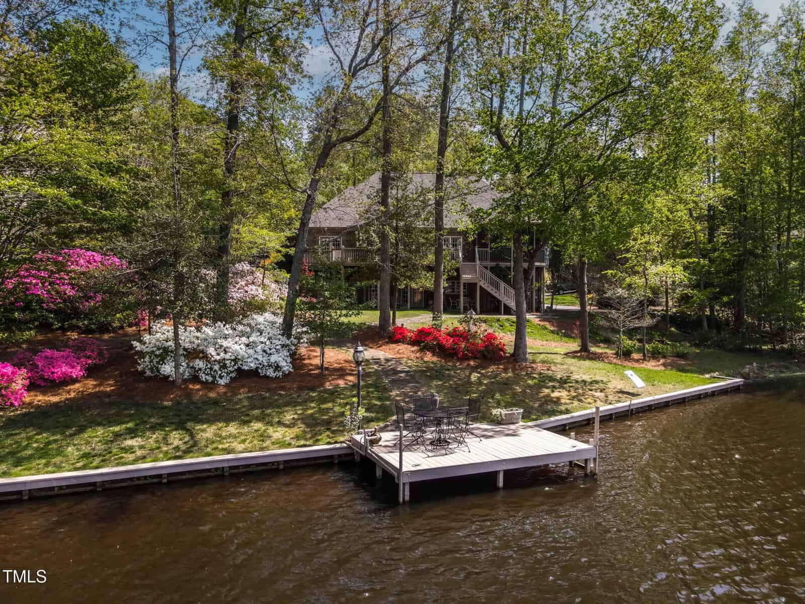 A wooden dock with chairs sits on a calm lake, facing a house surrounded by tall trees and blooming azalea bushes in pink, white, and red. The yard is grassy and partially shaded, with a wooded backdrop.