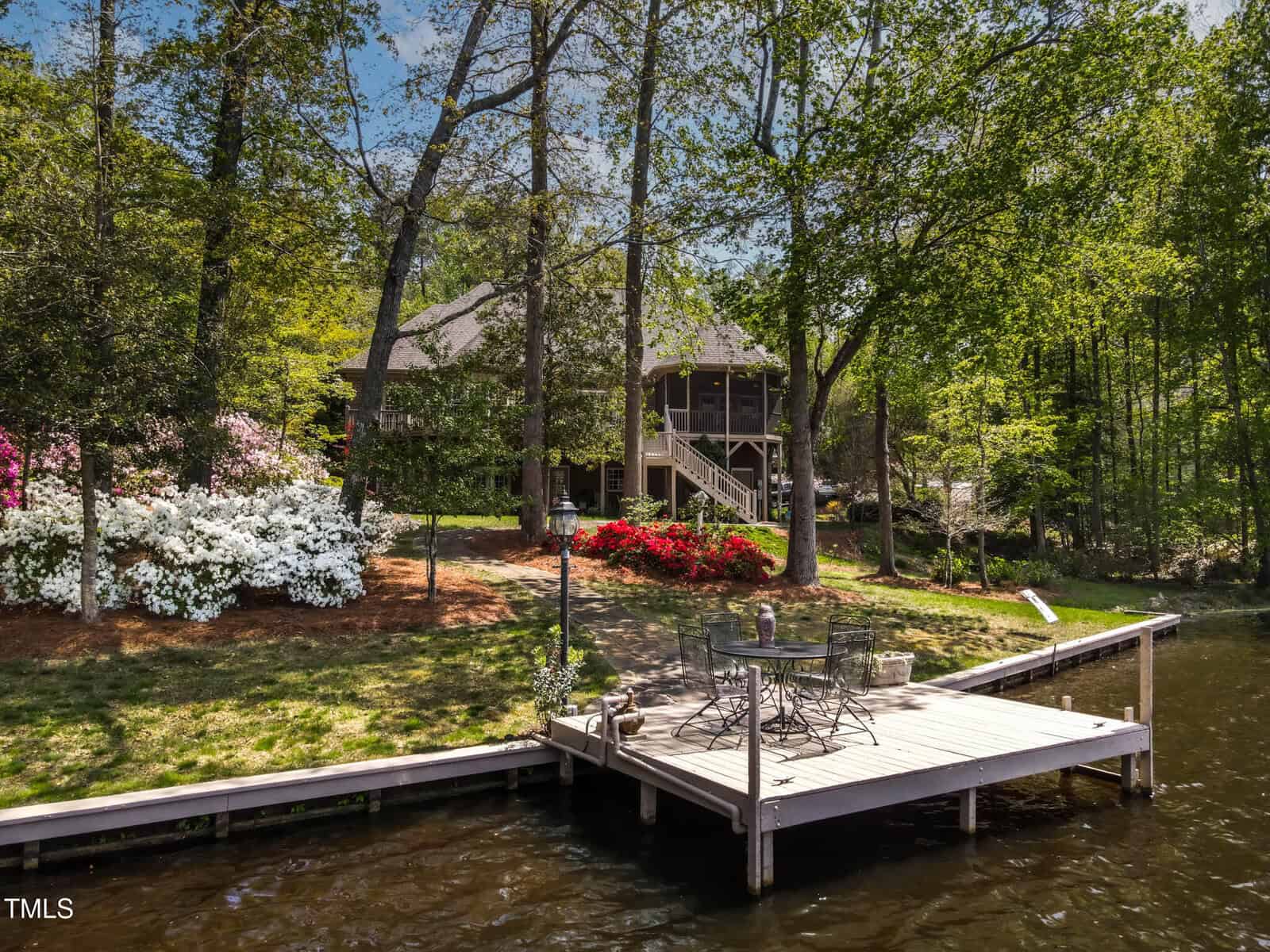 A wooden dock with a table and chairs extends over a lake, surrounded by lush trees and blooming colorful flowers, with a house visible in the background among the greenery.