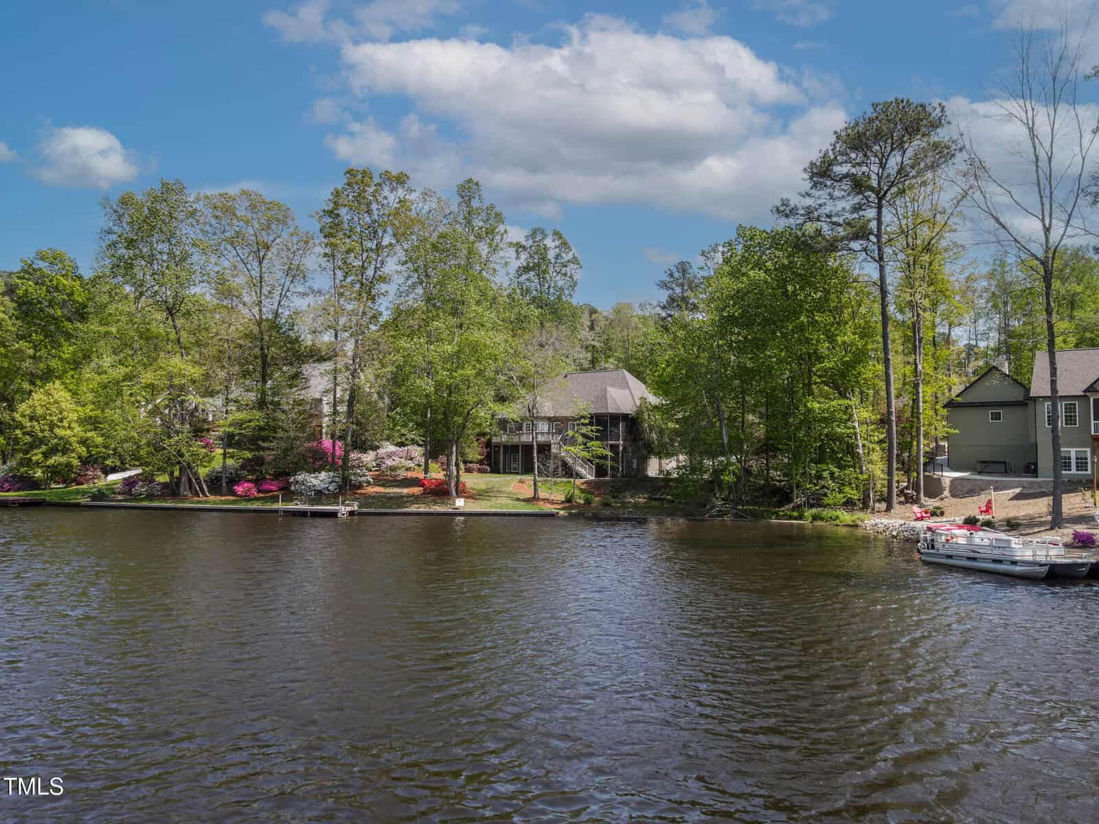 A lakefront view of houses surrounded by trees, with decks facing the water, colorful flowers along the shore, and boats docked near the edge on a partly cloudy day.
