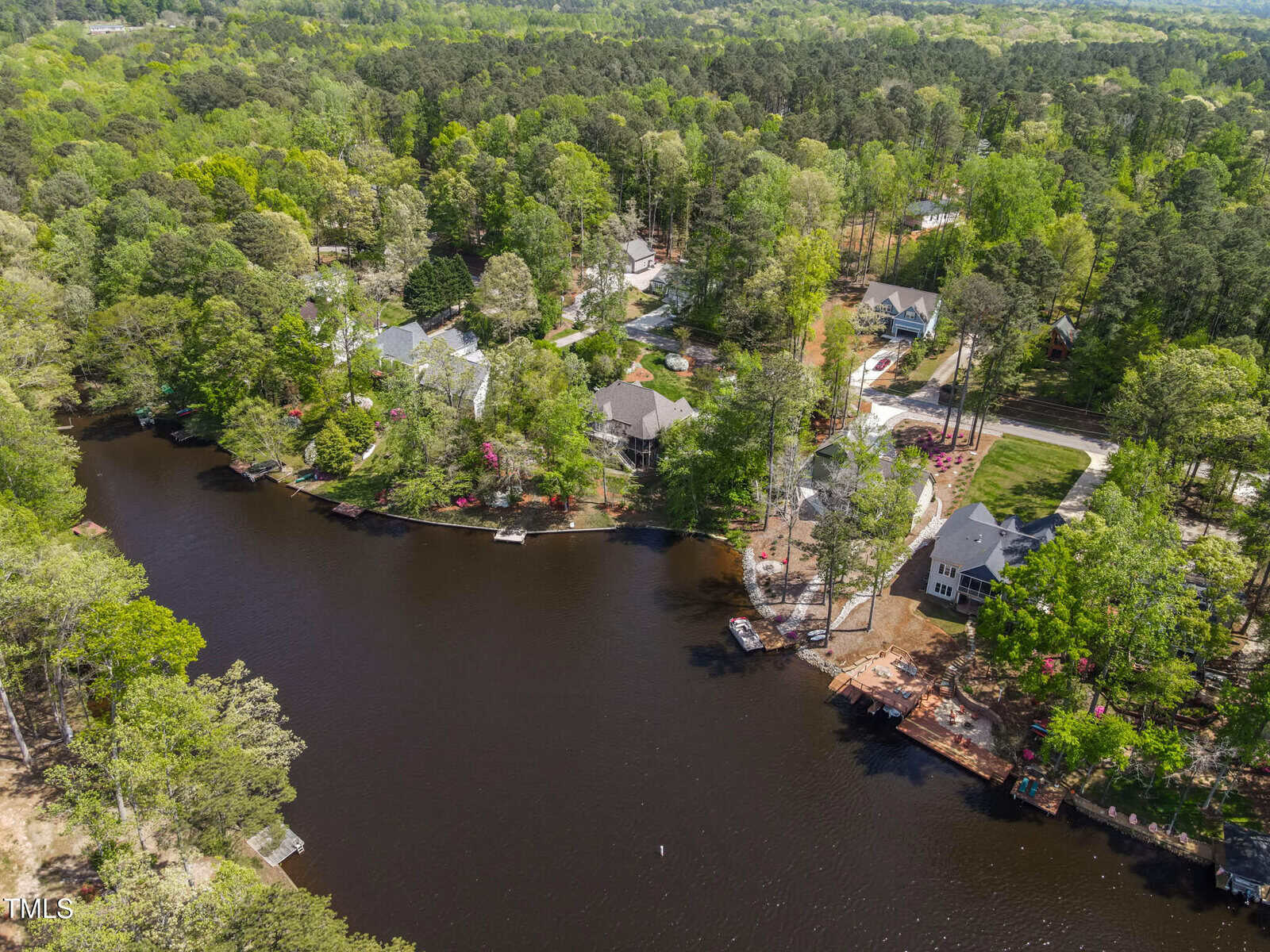 Aerial view of houses nestled among trees along the shoreline of a calm, winding lake in a green, wooded area on a sunny day.