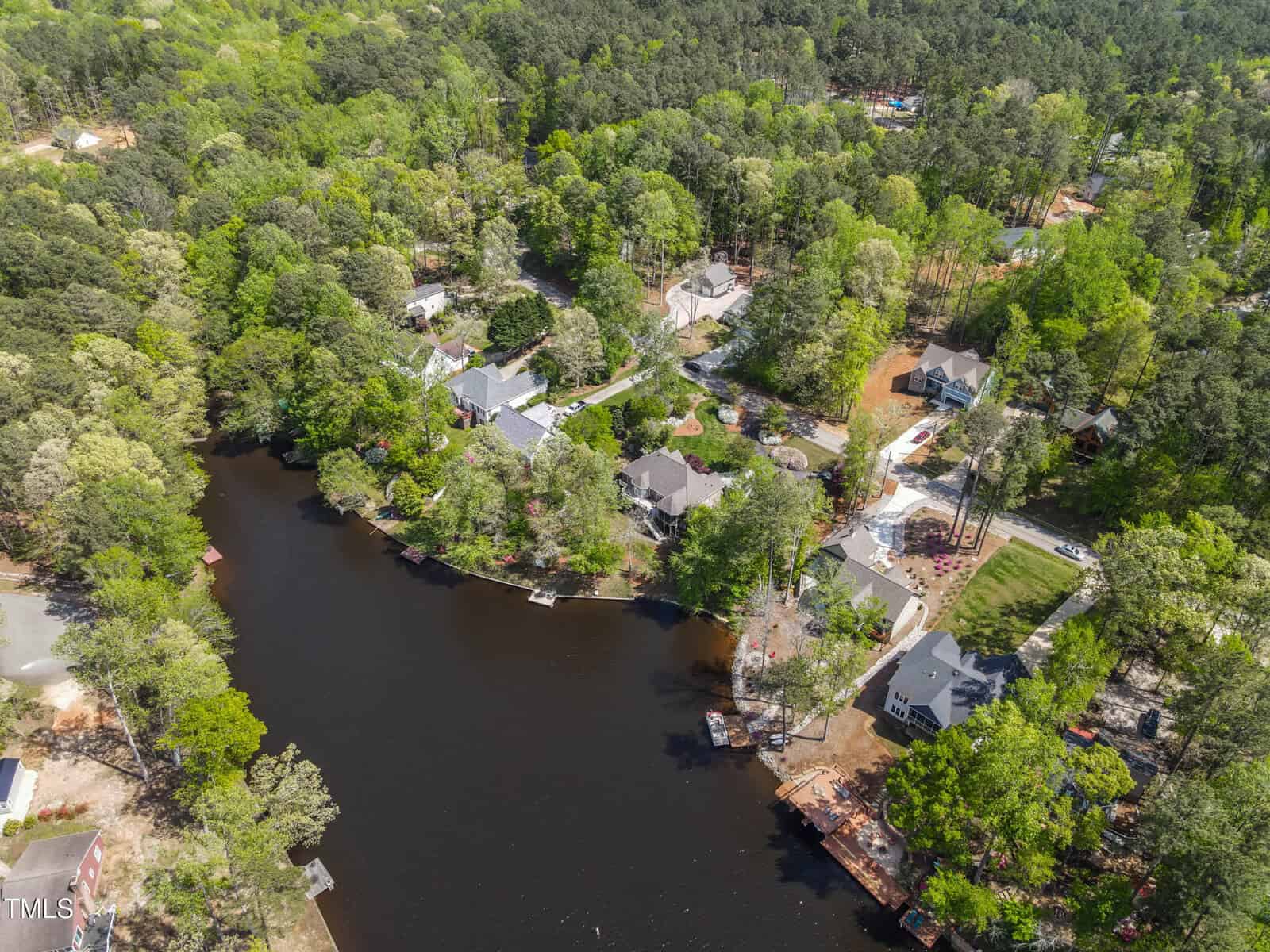 Aerial view of a wooded lakeside neighborhood with several houses, docks, and boats along the shoreline, surrounded by dense green trees and forested areas.