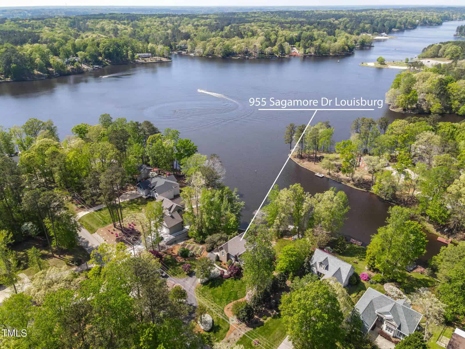 Aerial view of a lakeside neighborhood with trees and several houses. A labeled line points to a home at 955 Sagamore Dr, Louisburg, near the water. A boat is seen moving on the lake.