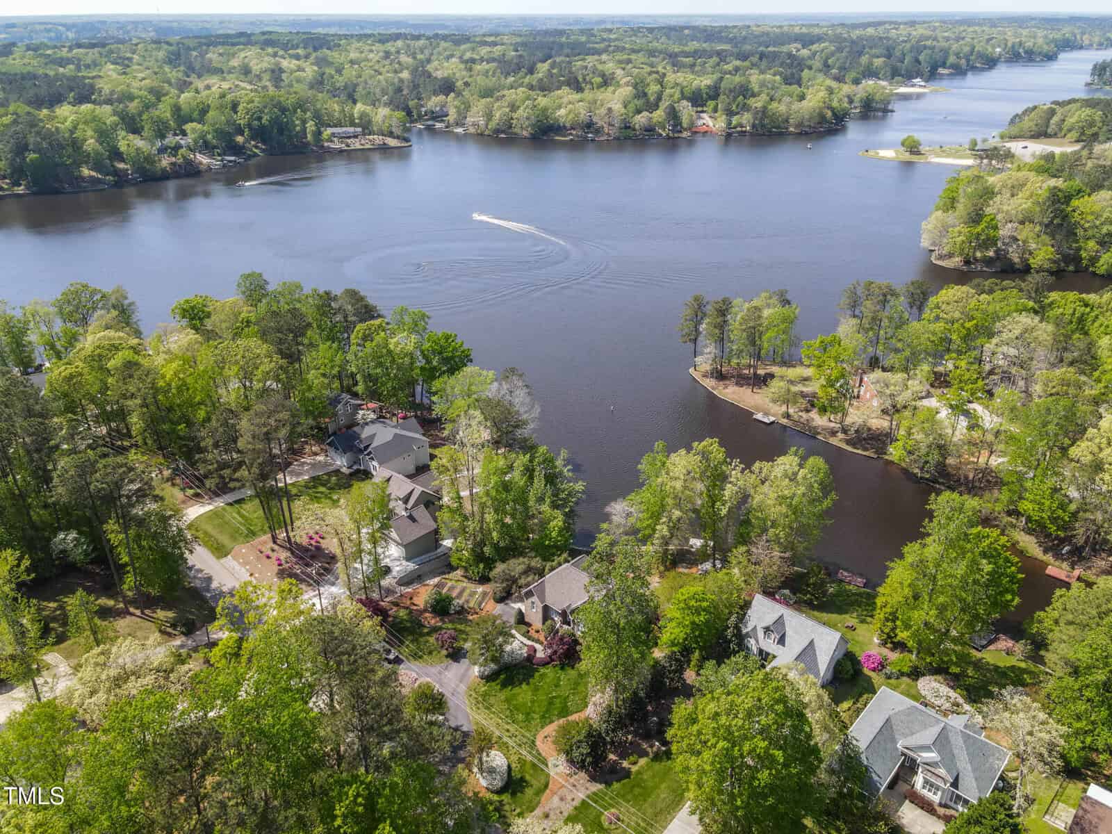 Aerial view of lakeside homes surrounded by trees, with a boat creating a wake on the water. The landscape features multiple peninsulas and a mix of greenery and houses along the shoreline under a clear sky.