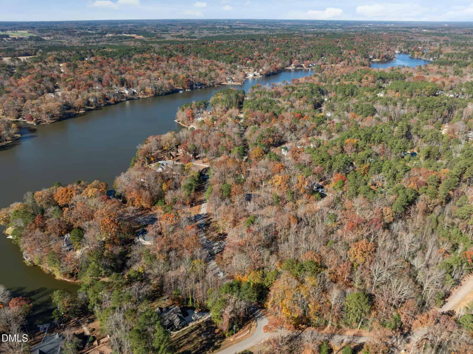 Aerial view of a wooded lakeside area in autumn, with houses scattered among colorful trees and winding roads near a large lake and inlets. The landscape extends to a distant horizon under a partly cloudy sky.