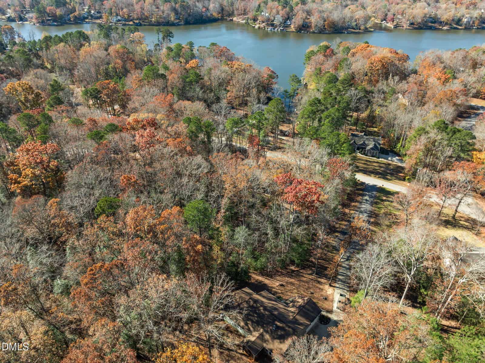 Aerial view of a wooded area with autumn foliage near a lake, scattered houses, and surrounding trees, with calm water in the background and a mix of orange, red, and green hues.