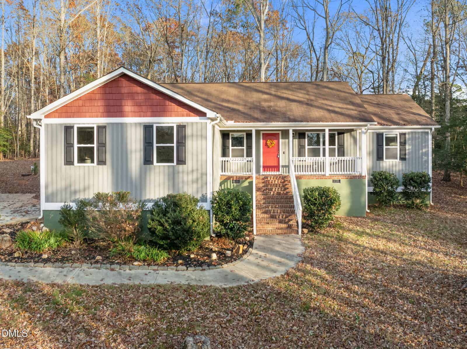A single-story house with light gray siding, red gable accents, a covered front porch with white railings, a red front door, brick steps, and surrounded by trees and shrubs.