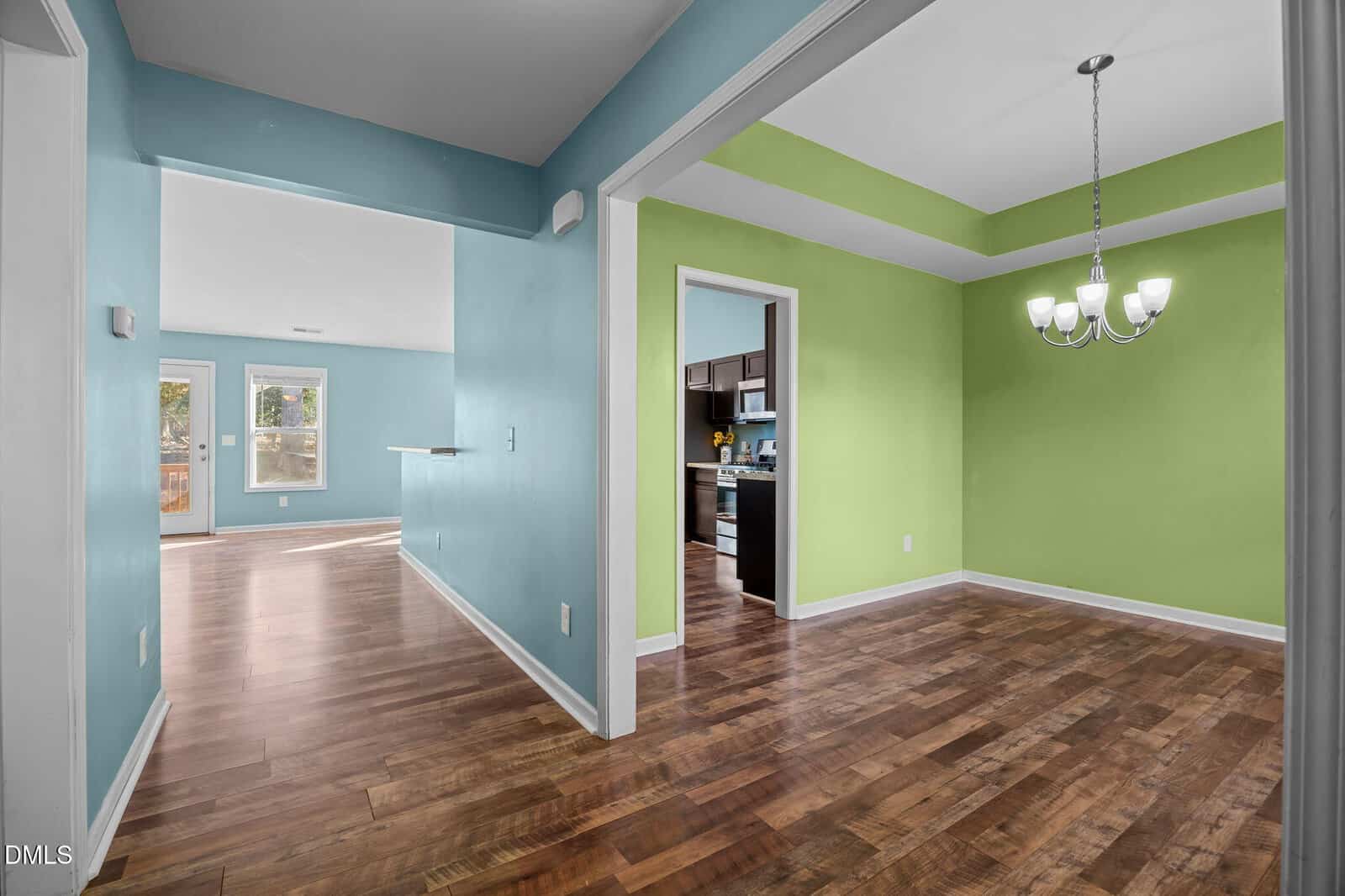 Interior view of a house with teal and lime green walls, hardwood floors, and a dining area featuring a modern chandelier. The kitchen and living area are visible through doorways, with natural light coming in from glass doors.