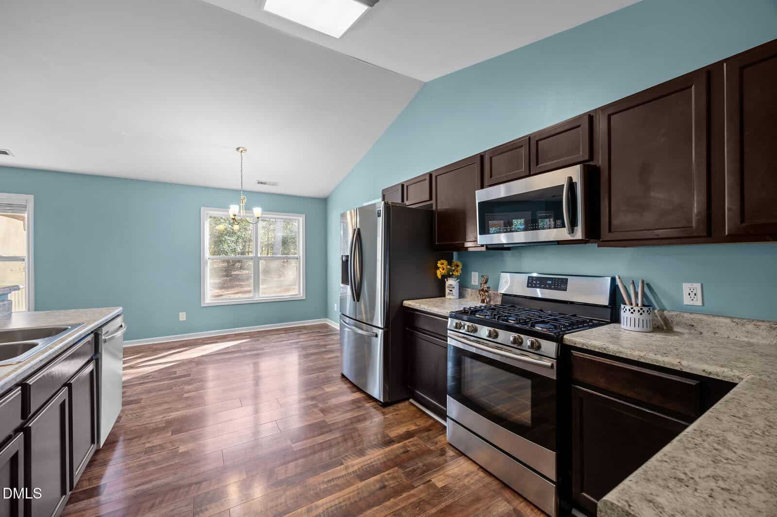 Modern kitchen with dark wood cabinets, stainless steel appliances, marble countertops, and blue walls. Large window lets in natural light, and there is a dining area with a chandelier in the background.