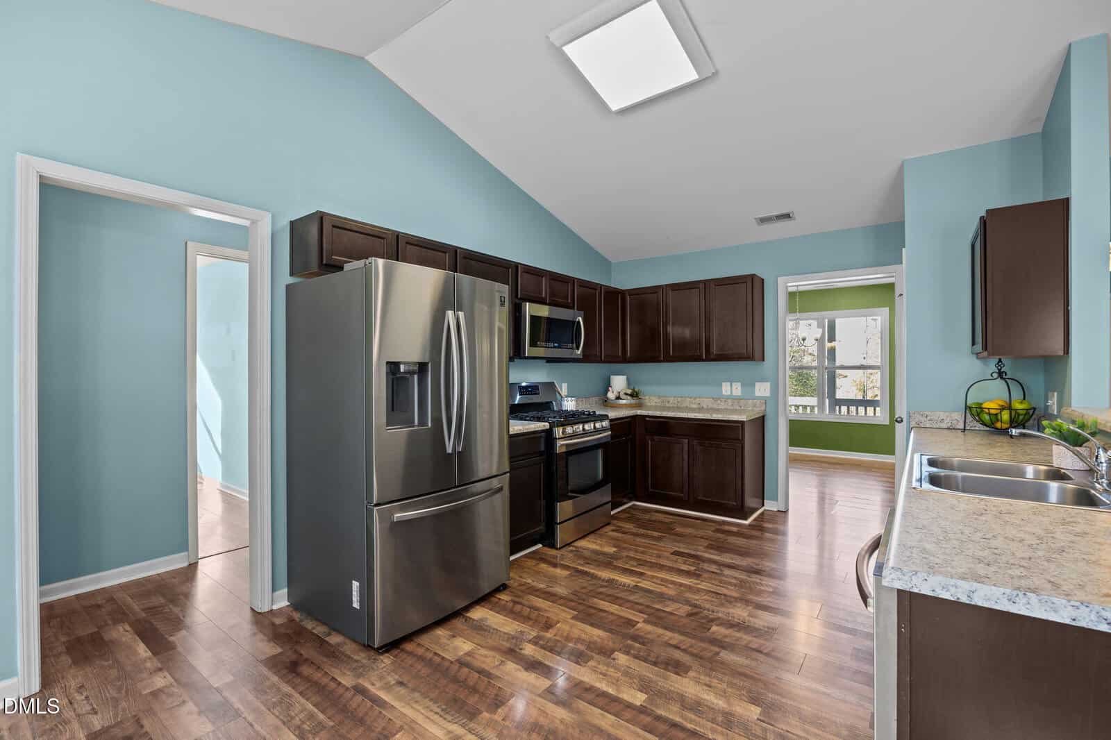 Modern kitchen with stainless steel appliances, dark wood cabinets, and marble countertops. The walls are painted light blue and the floor is wooden. Two doorways lead to adjacent rooms, one with green walls.