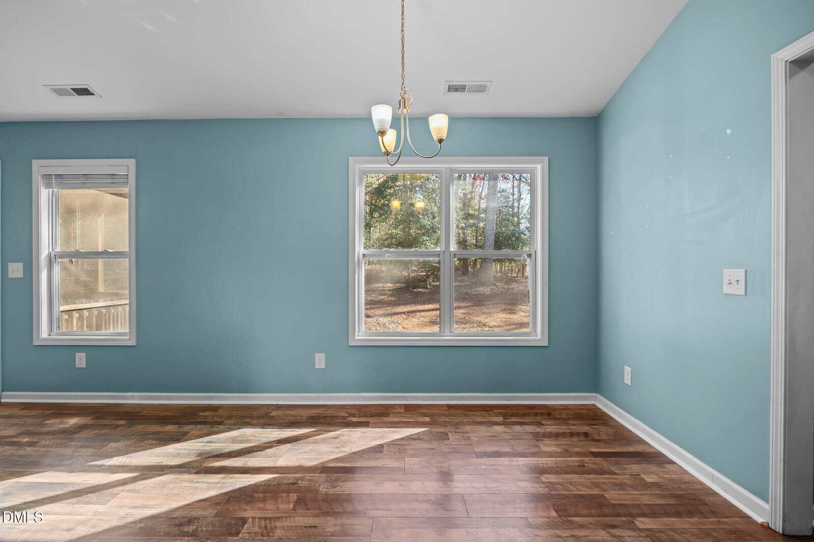 A room with blue walls, two large windows, a hanging light fixture, and wooden floors. Sunlight streams through the windows, casting shadows on the floor. The room appears empty and well-lit.